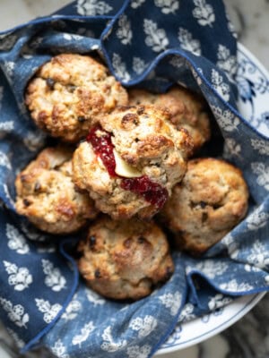 A bowl lined with blue and white patterned cloth holds several golden brown Irish soda bread scones, one split open and topped with butter and red jam, just like Mary O's copycat recipe—perfect with currants for a classic touch.