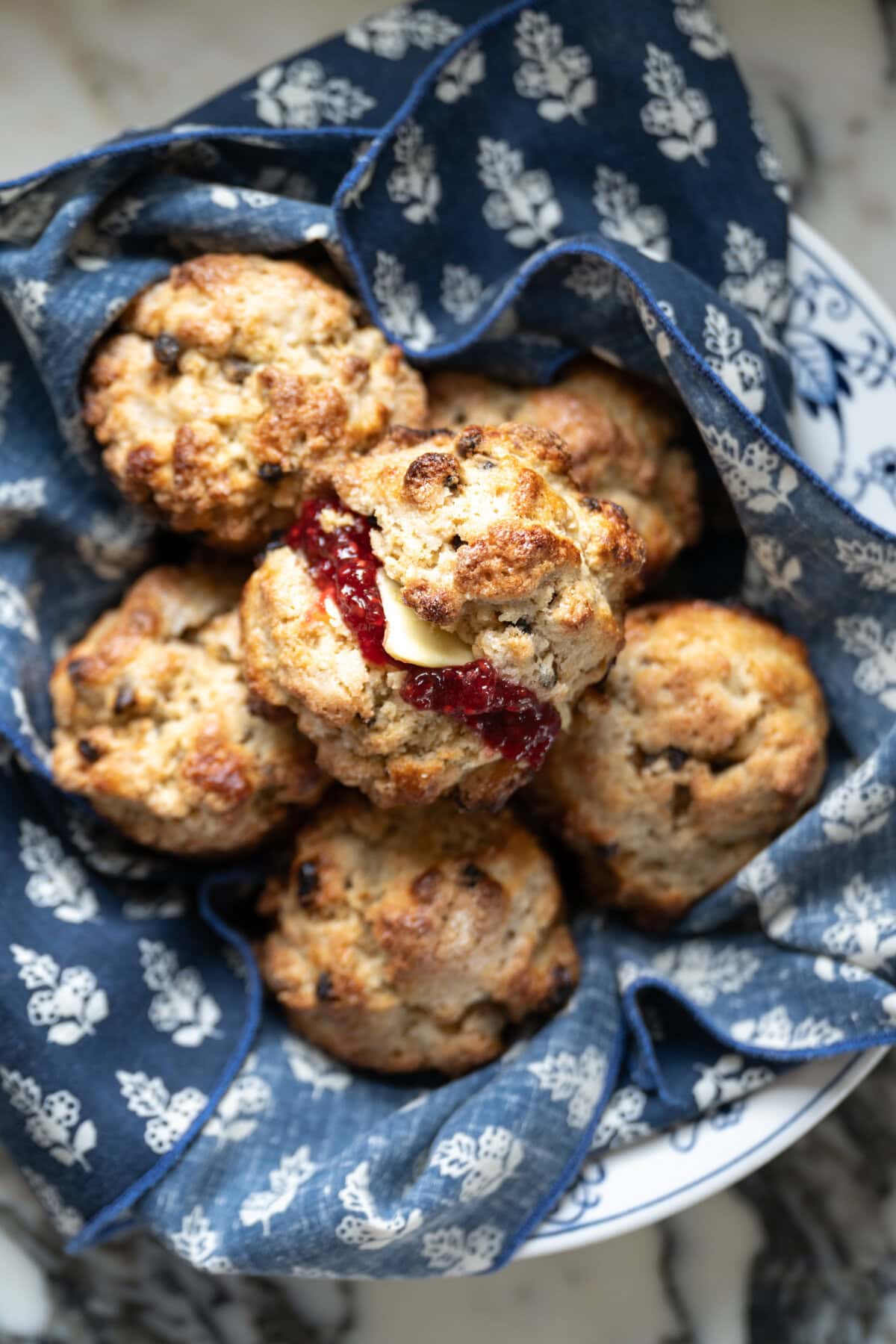 A bowl lined with blue and white patterned cloth holds several golden brown Irish soda bread scones, one split open and topped with butter and red jam, just like Mary O's copycat recipe—perfect with currants for a classic touch.