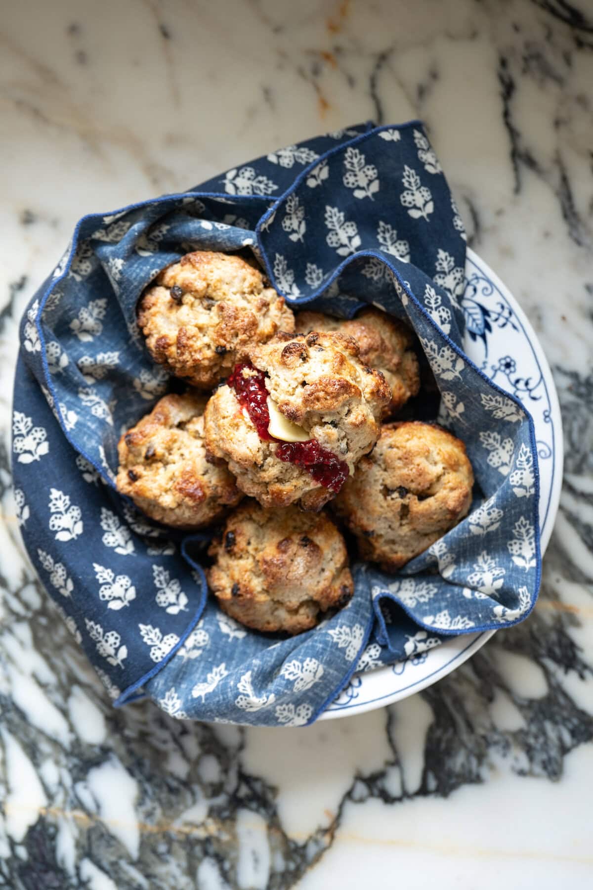 A bowl lined with a blue patterned cloth holds several rustic Irish soda bread scones, one of which is split open and spread with butter and jam, set on a marble surface.