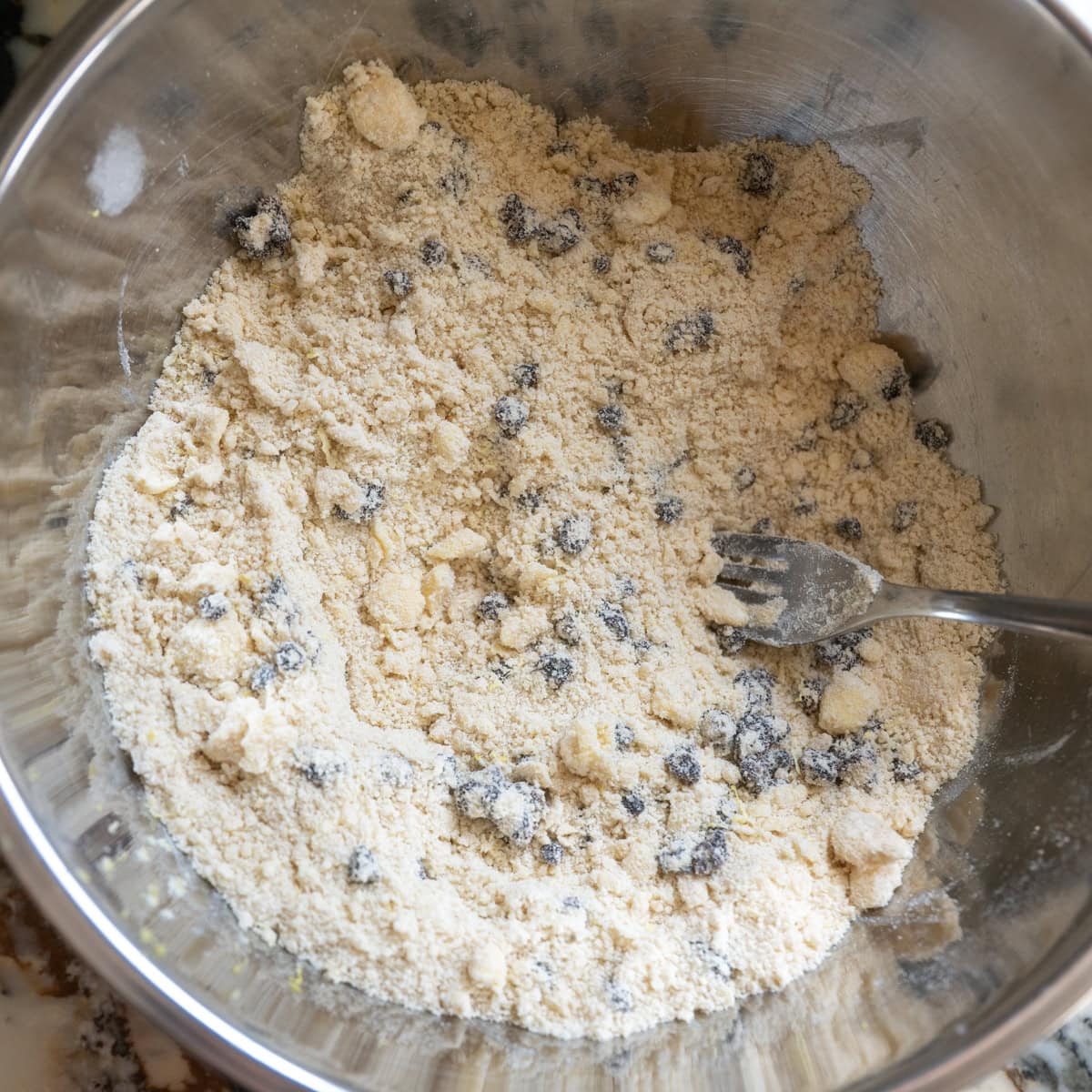 A metal mixing bowl filled with pale flour mixture and scattered dark currants, with a metal fork resting inside, suggesting Irish soda bread scone recipe preparation.