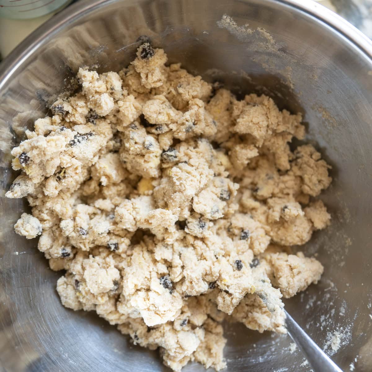 A metal mixing bowl filled with chunky Irish soda bread scone dough containing currants, with a spoon partially buried in the mixture.