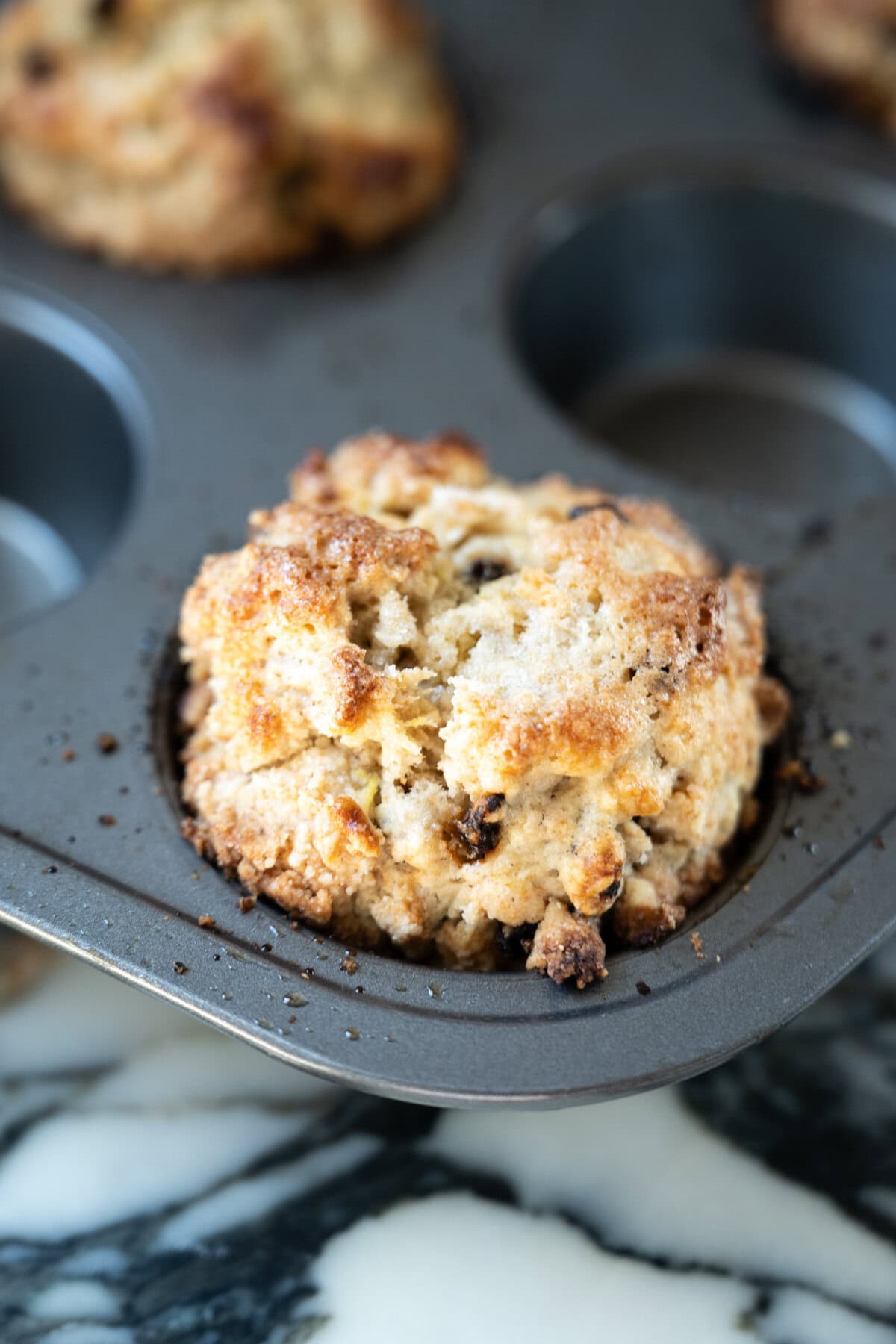 A close-up of a golden-brown Irish soda bread scone with a crumbly top sits in a metal muffin tin, with another scone partially visible in the background on a marbled surface.