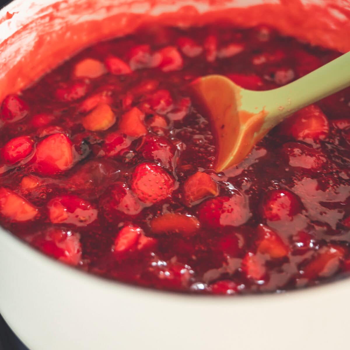 Strawberry rhubarb fruit filling cooking in a white saucepan with a green silicone spatula.