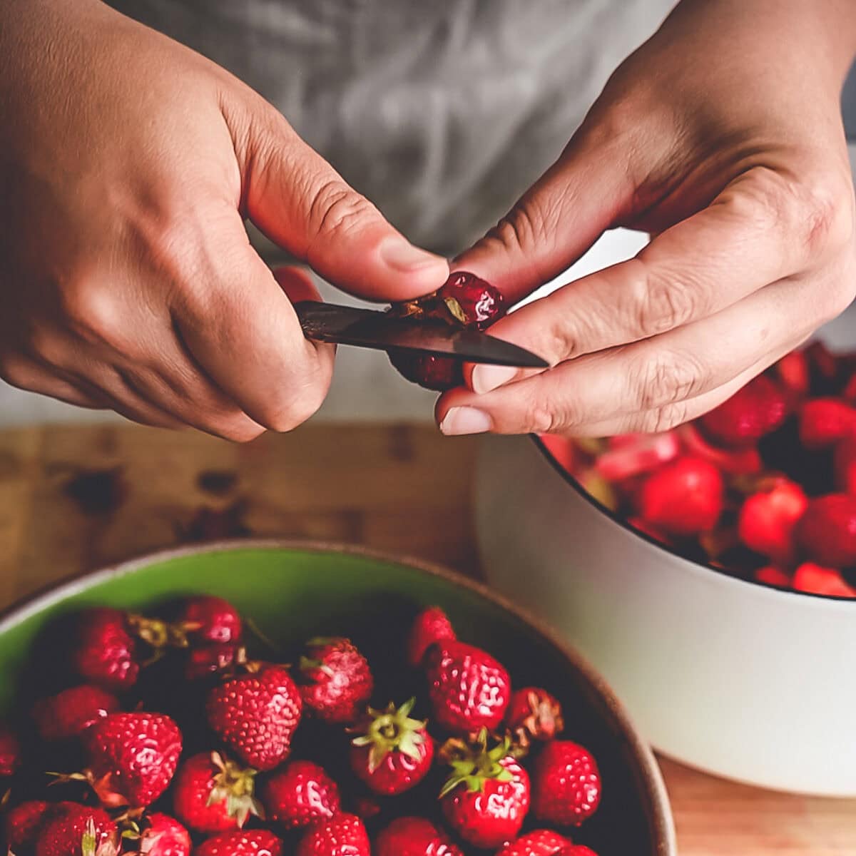 Removing the leafy top from a strawberry with a paring knife from a bowl full of strawberries.