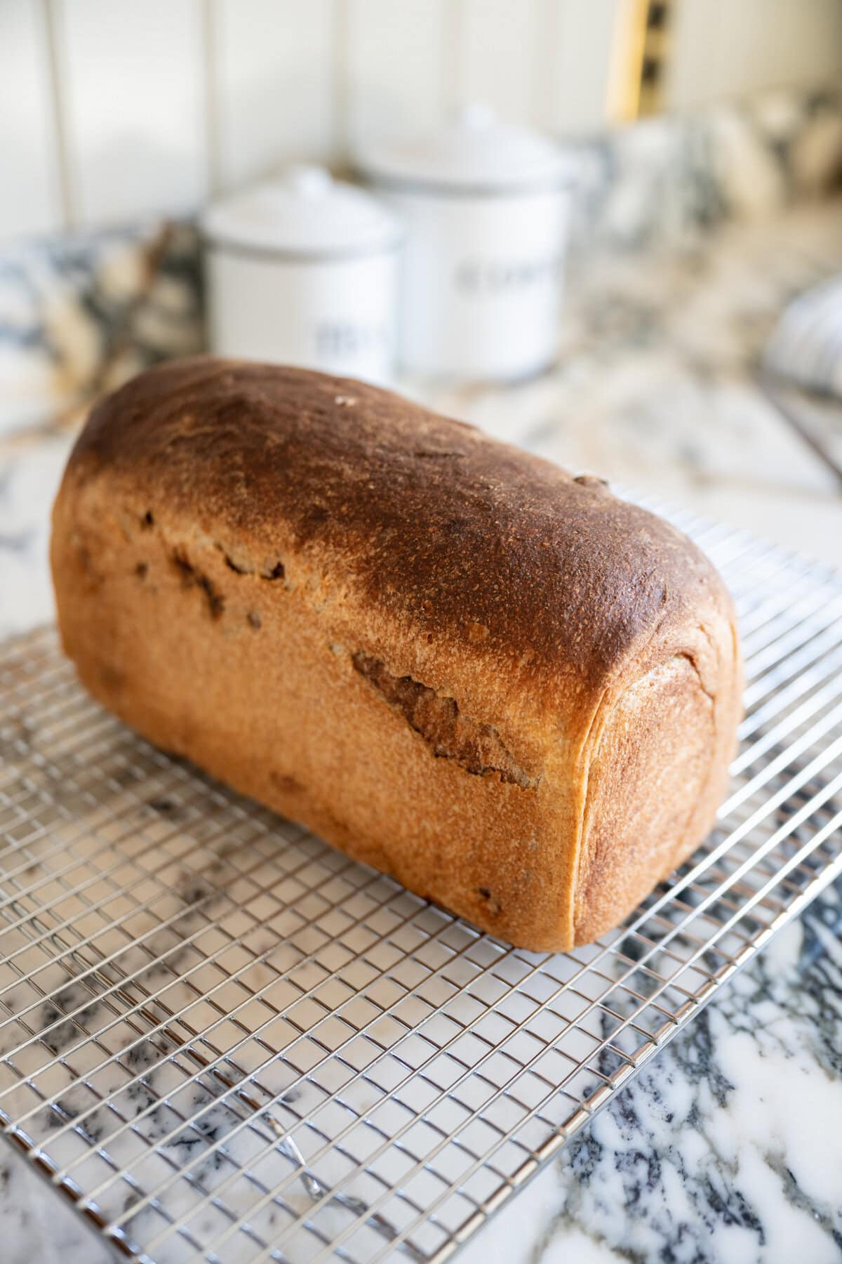 A golden-brown loaf of soft cinnamon raisin sourdough bread sits on a wire cooling rack, placed on a marble countertop.