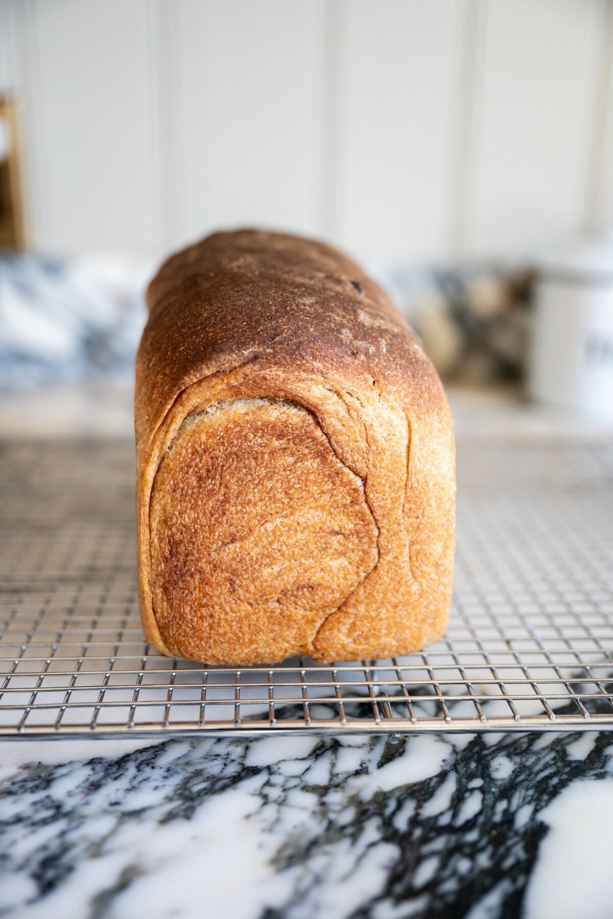 A finished loaf of golden-brown cinnamon raisin sourdough sandwich bread sits on a cooling rack atop a marble countertop, with a blurred kitchen background.