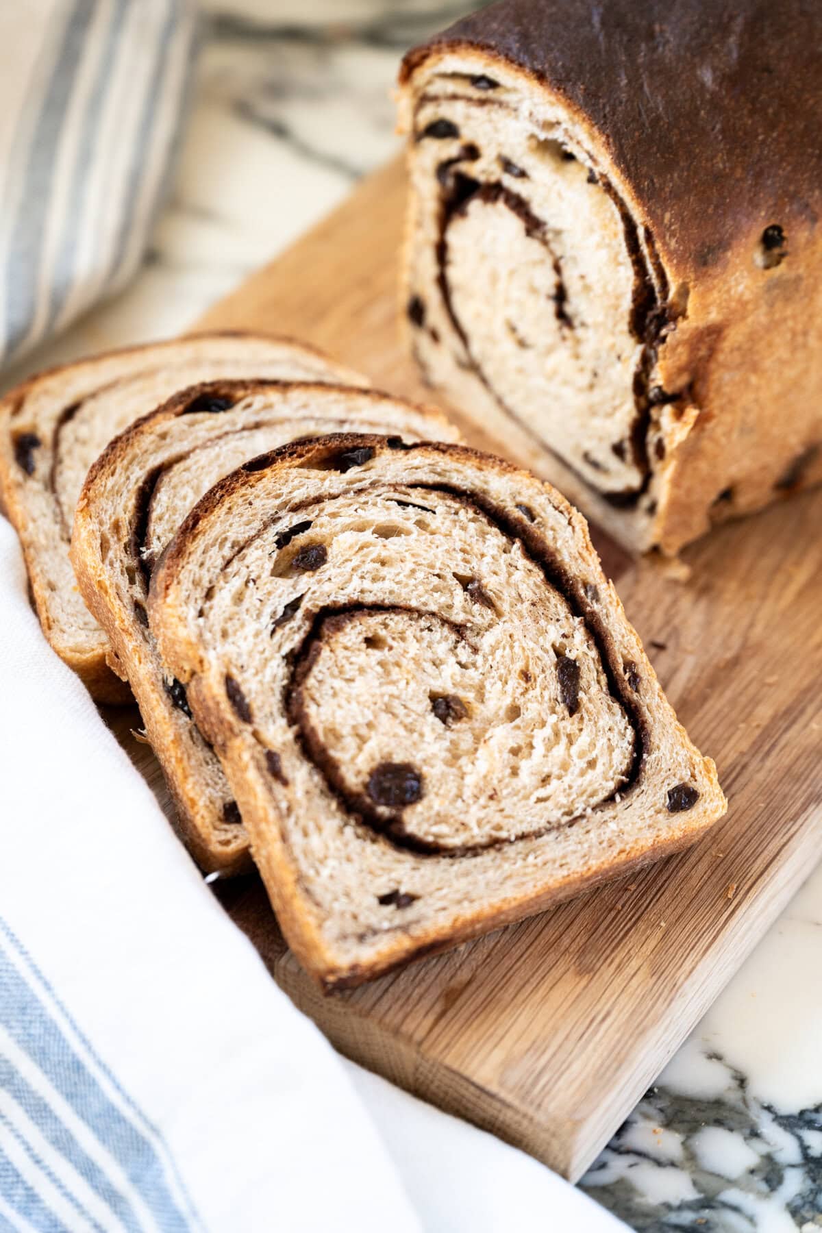 Sliced cinnamon raisin sourdough sandwich loaf on a wooden cutting board, with a white and blue striped cloth nearby on a marble surface.
