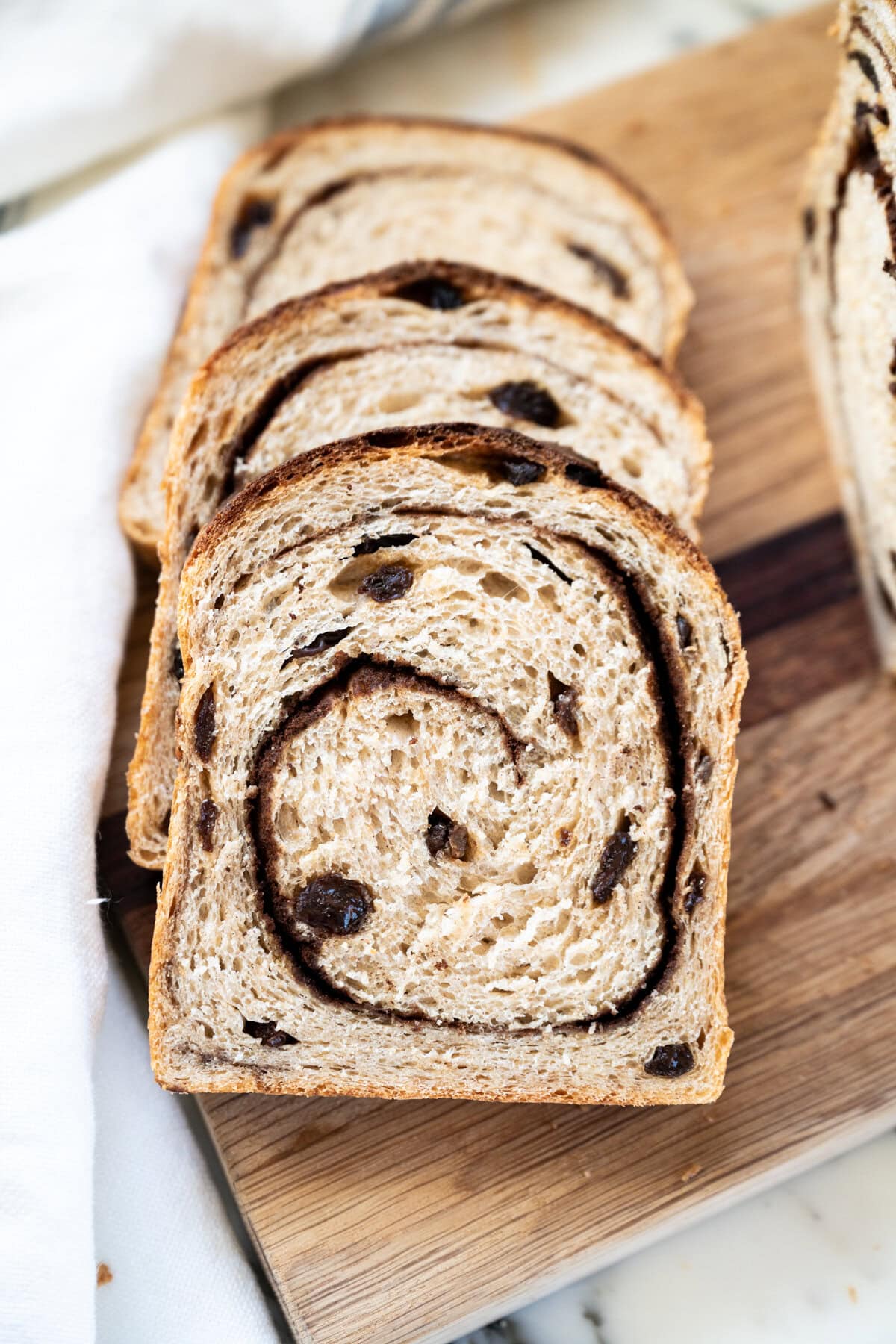 Four slices of Cinnamon raisin sourdough sandwich bread with a visible swirl pattern and raisins are arranged on a wooden cutting board. A white cloth and the remaining loaf are partially visible in the background.