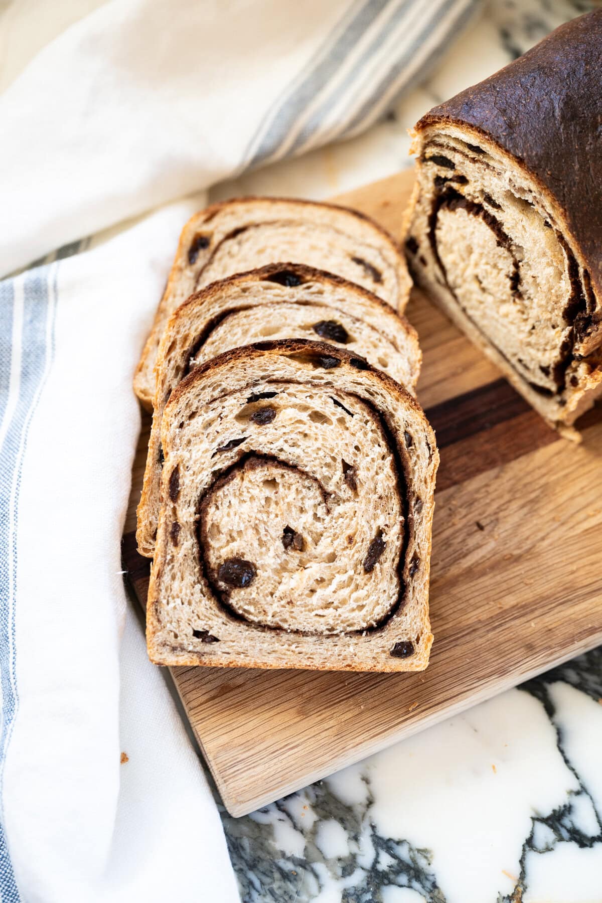 Three slices of cinnamon raisin sourdough bread with a visible swirl and raisins, arranged on a wooden cutting board next to a loaf. A white towel with blue stripes is nearby on a marble surface.