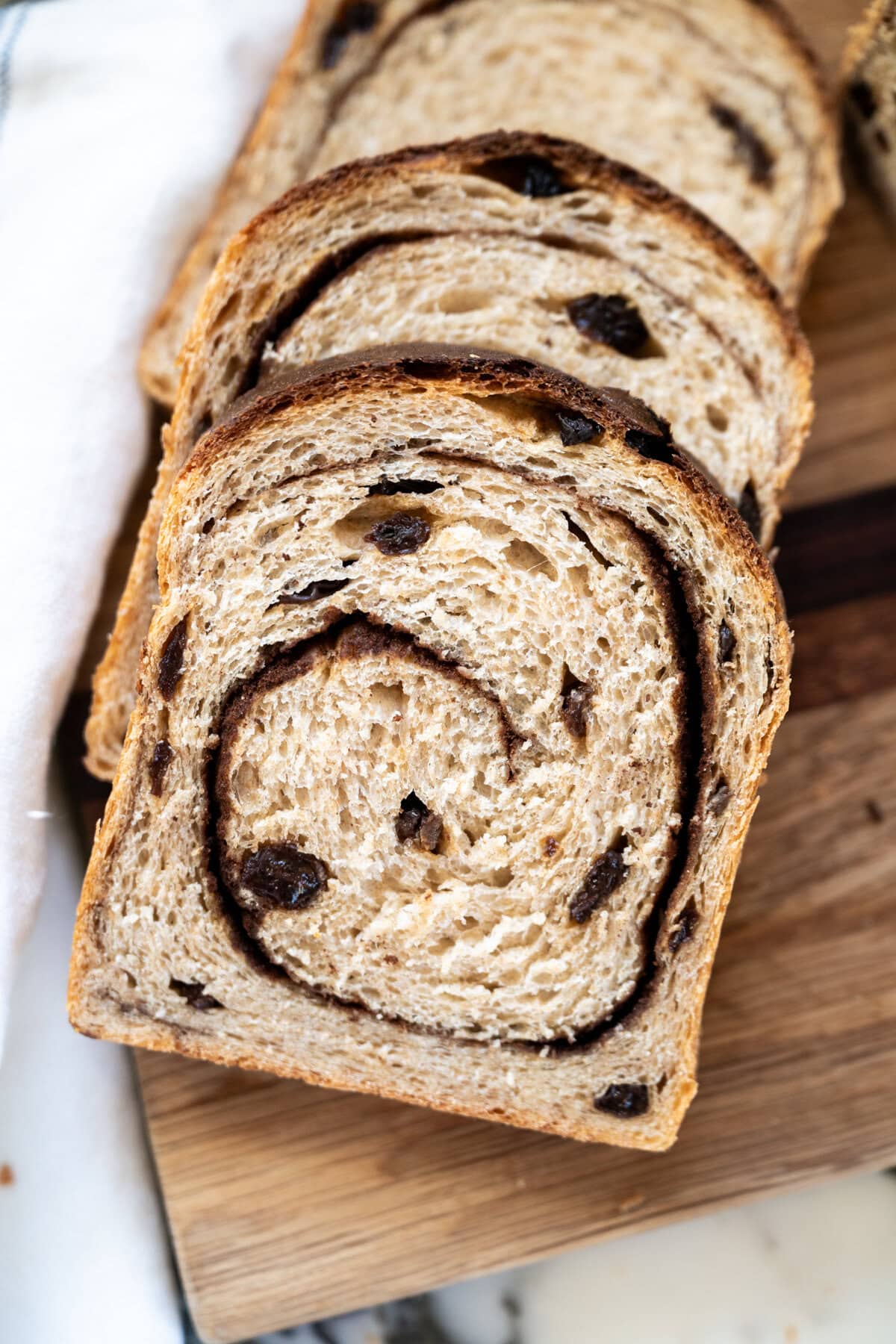 Three slices of cinnamon raisin sourdough sandwich bread are arranged on a wooden cutting board, showing a swirl of cinnamon and scattered raisins throughout the soft, textured bread.