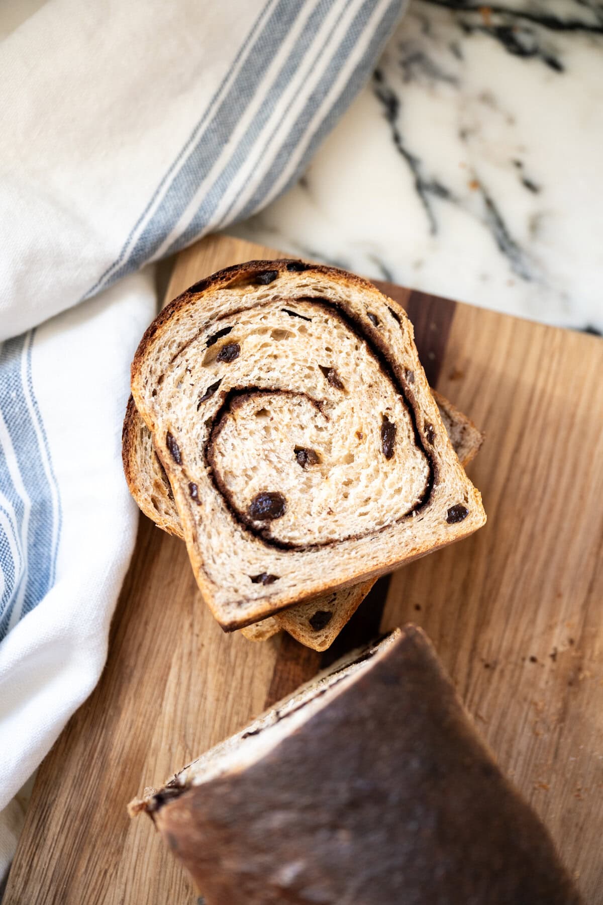 Slices of homemade cinnamon raisin sourdough bread sit on a wooden cutting board next to a loaf, with a white and blue striped kitchen towel nearby on a marble countertop.