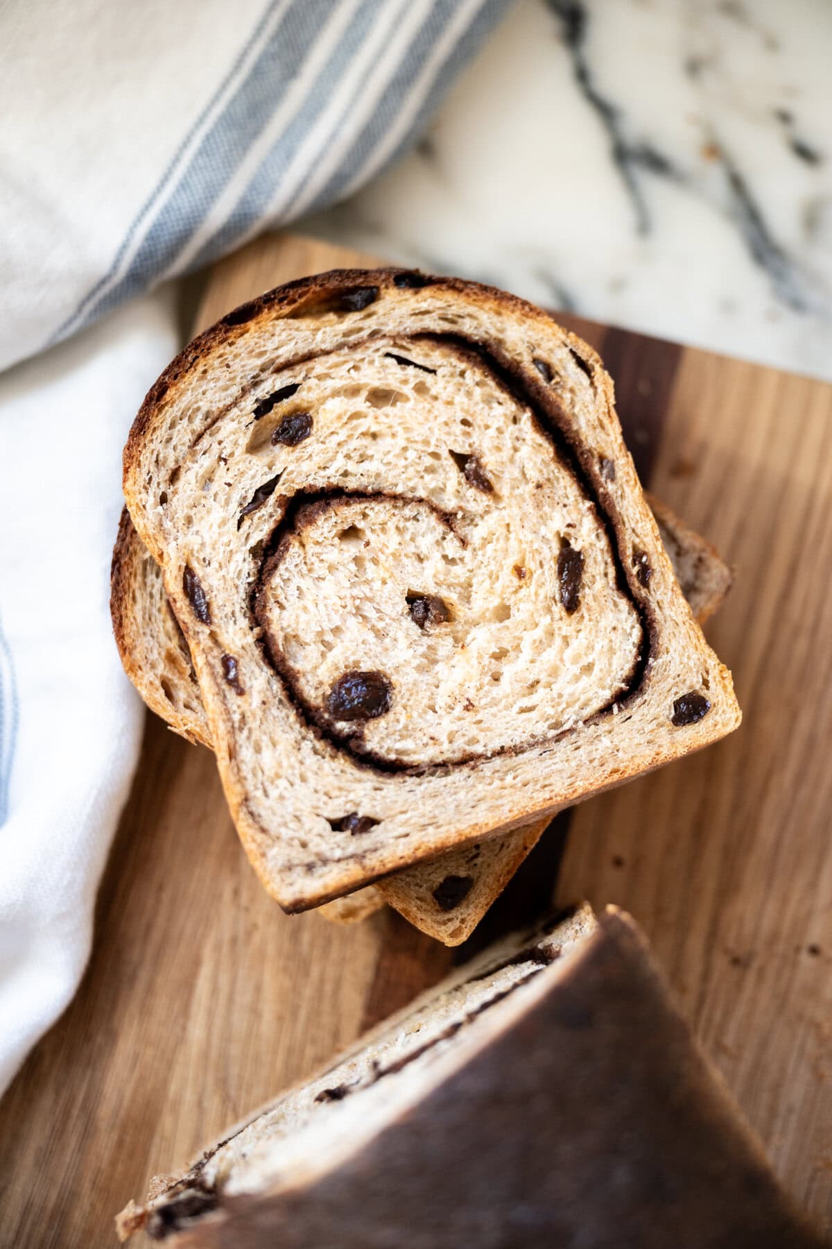 Close-up stacked slices of cinnamon raisin sourdough bread with raisins on a wooden surface, with part of a loaf and a white towel with blue stripes visible nearby.