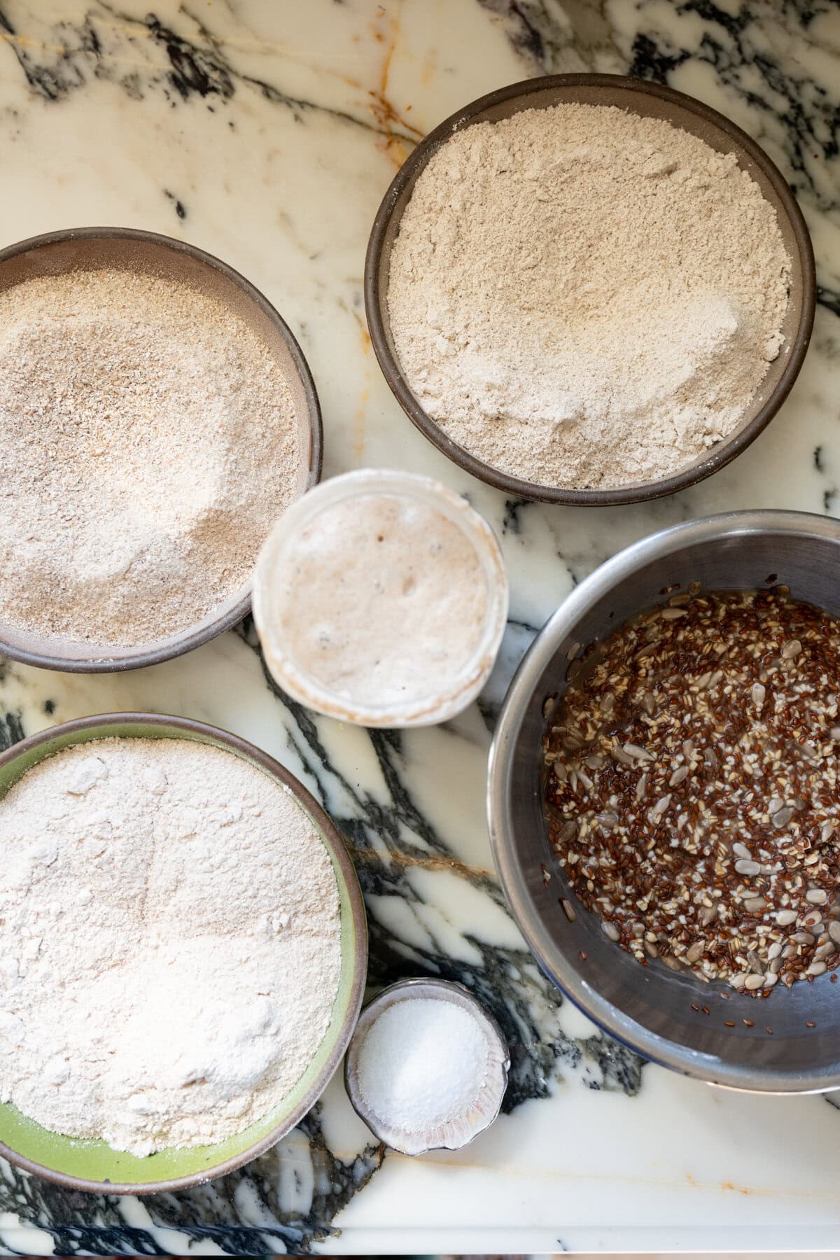 Ingredients for Danish rye bread rugbrød including dark rye flour, whole wheat flour, salt, cracked rye berries, sourdough starter, and presoaked steel cut oats, sunflower seeds, and flax seeds.