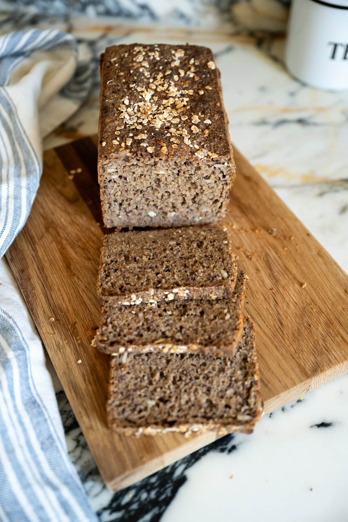 Homemade Danish rye bread loaf with oats on top sits on a wooden cutting board, partially sliced, with three slices fanned out in front. A striped cloth is nearby on a marble countertop.