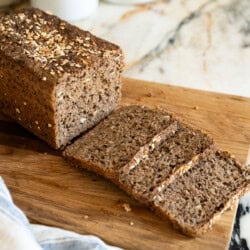 A loaf of brown, seeded Danish rye bread is partially sliced on a wooden cutting board, with a kitchen towel nearby and white containers labeled "TEA" and "COFFEE" in the background.