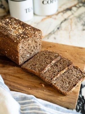 A loaf of brown, seeded Danish rye bread is partially sliced on a wooden cutting board, with a kitchen towel nearby and white containers labeled "TEA" and "COFFEE" in the background.