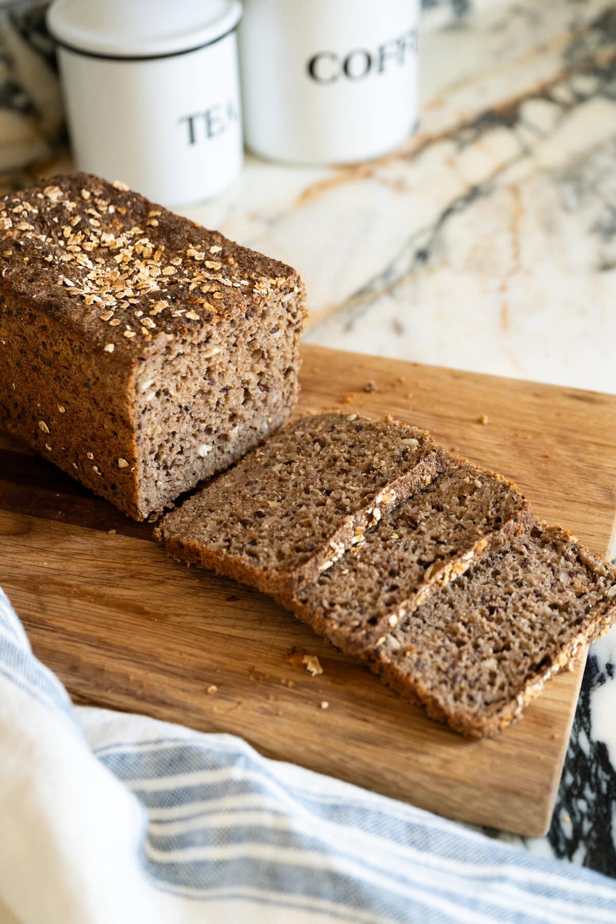 A loaf of Easy sourdough Danish rye bread recipe (rugbrød) with seeds, sliced and ready to serve on a wooden cutting board, with a kitchen towel nearby and white containers labeled "TEA" and "COFFEE" in the background.