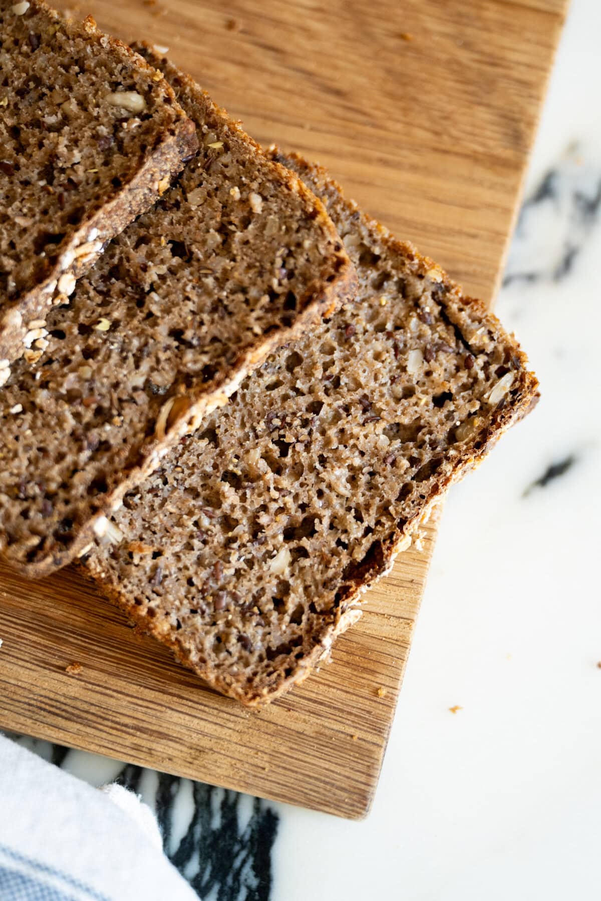 Three slices of dense, whole grain Danish rye bread rugbrød cut for open-faced sandwiches rest on a wooden cutting board, set on a white and black marble surface.