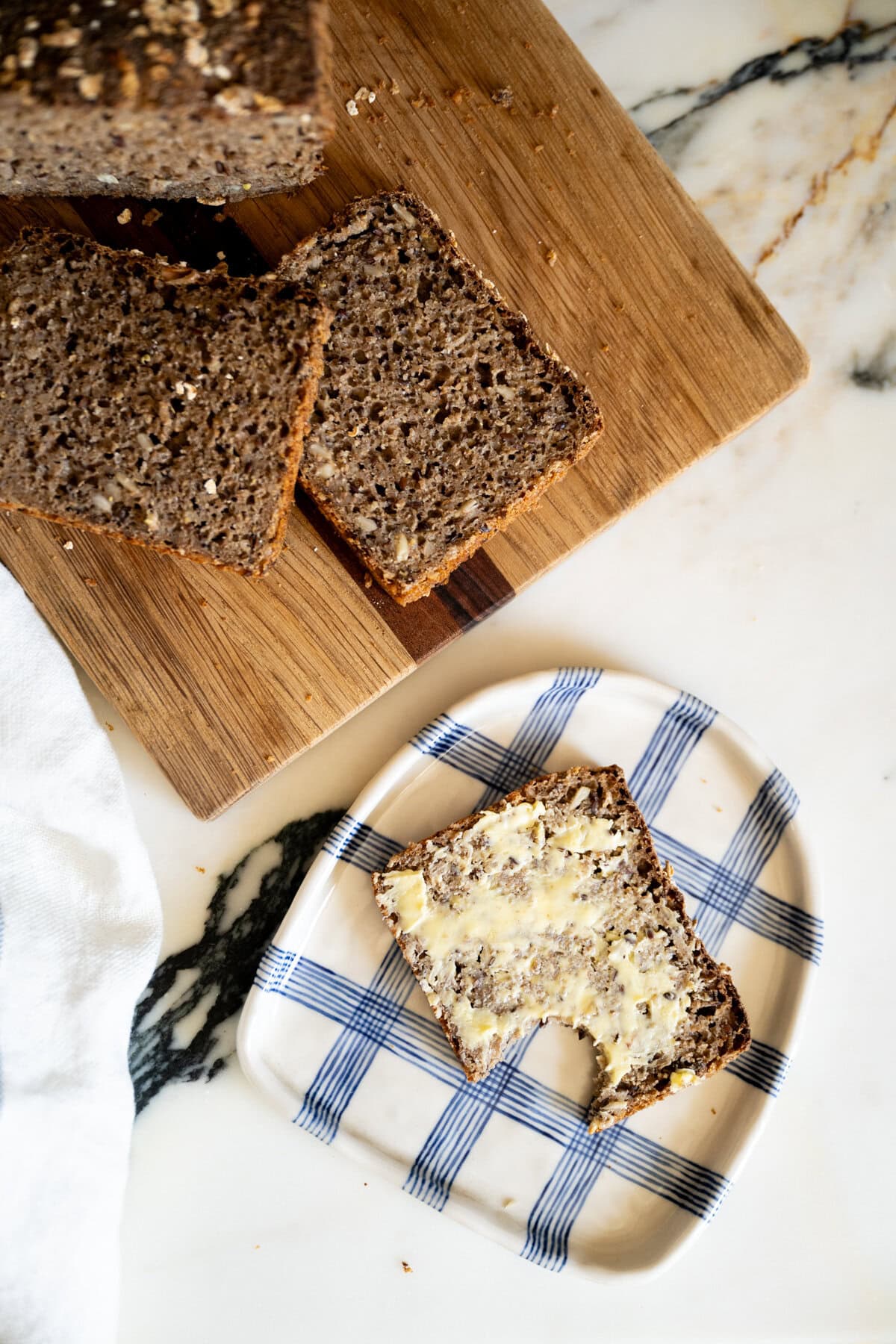 A slice of whole grain rugbrød - dense Danish rye bread - with butter and a bite taken out sits on a checkered plate beside a cutting board with two more slices and a loaf on a marble surface.
