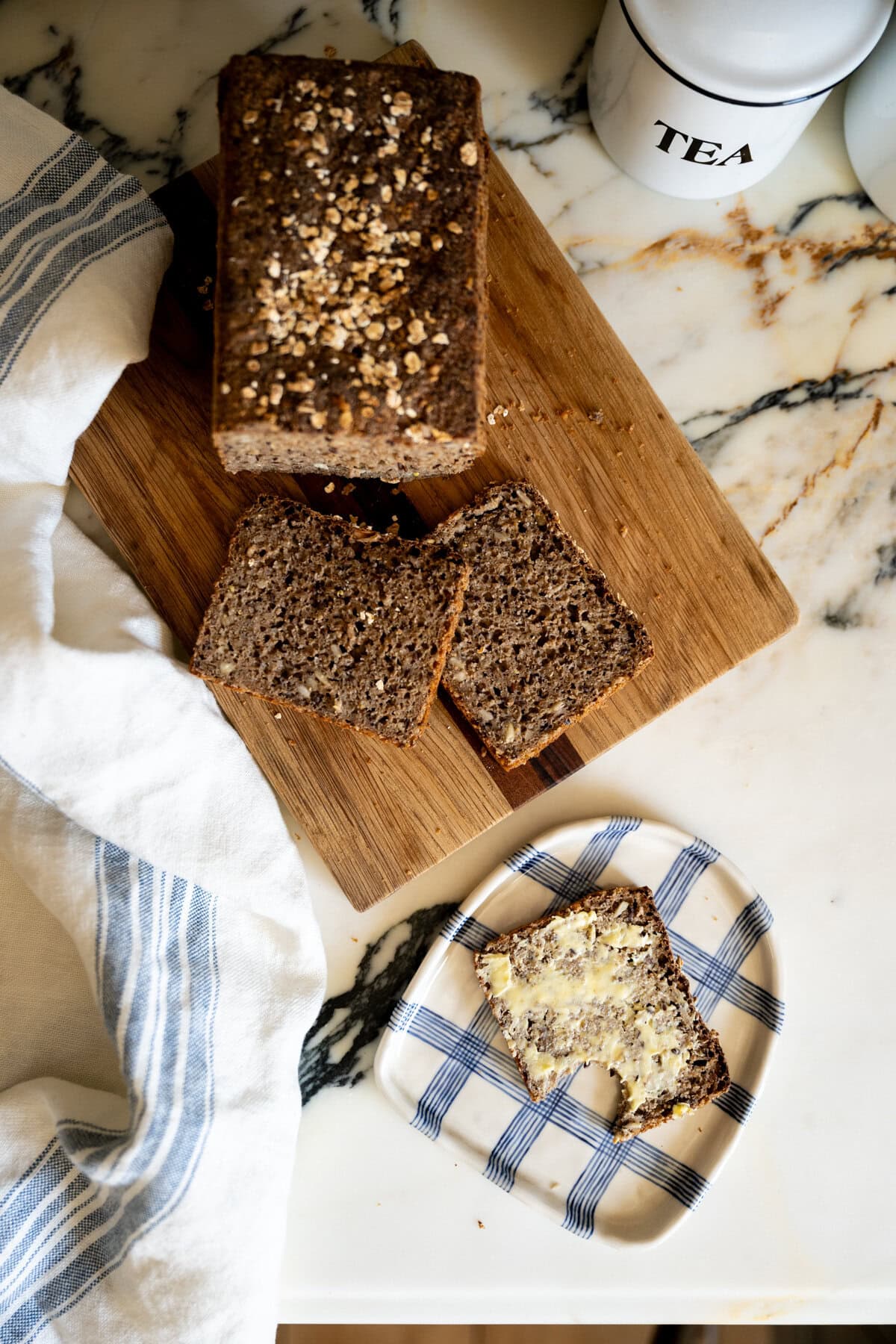 A loaf of seeded Danish rye bread (rugbrød) on a wooden cutting board, with two slices cut and one slice on a checkered plate, spread with butter. A striped kitchen towel and a tea container are nearby on a marble countertop.