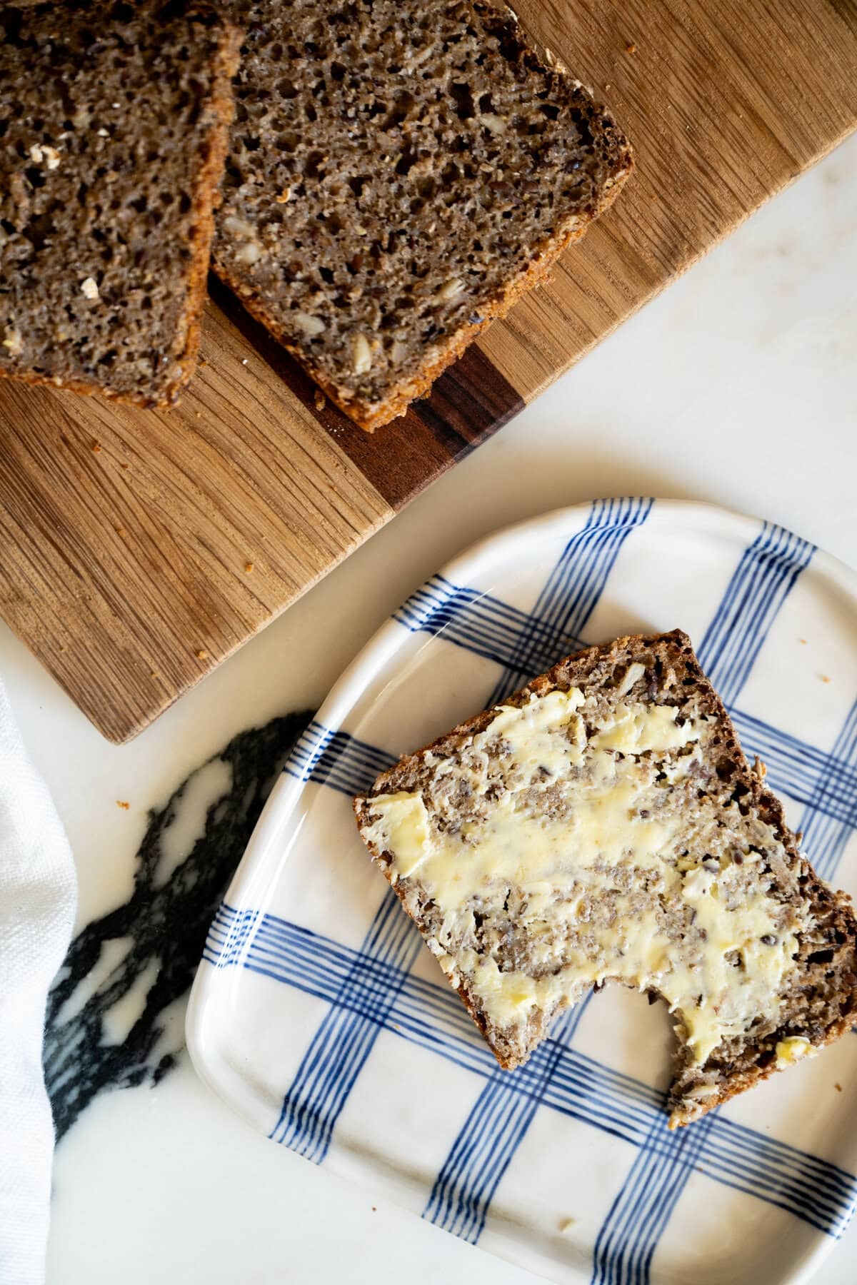 A slice of Danish rugbrød rye bread with butter and a bite taken out sits on a blue plaid plate, next to a cutting board with more slices of bread on a white and black marble surface.