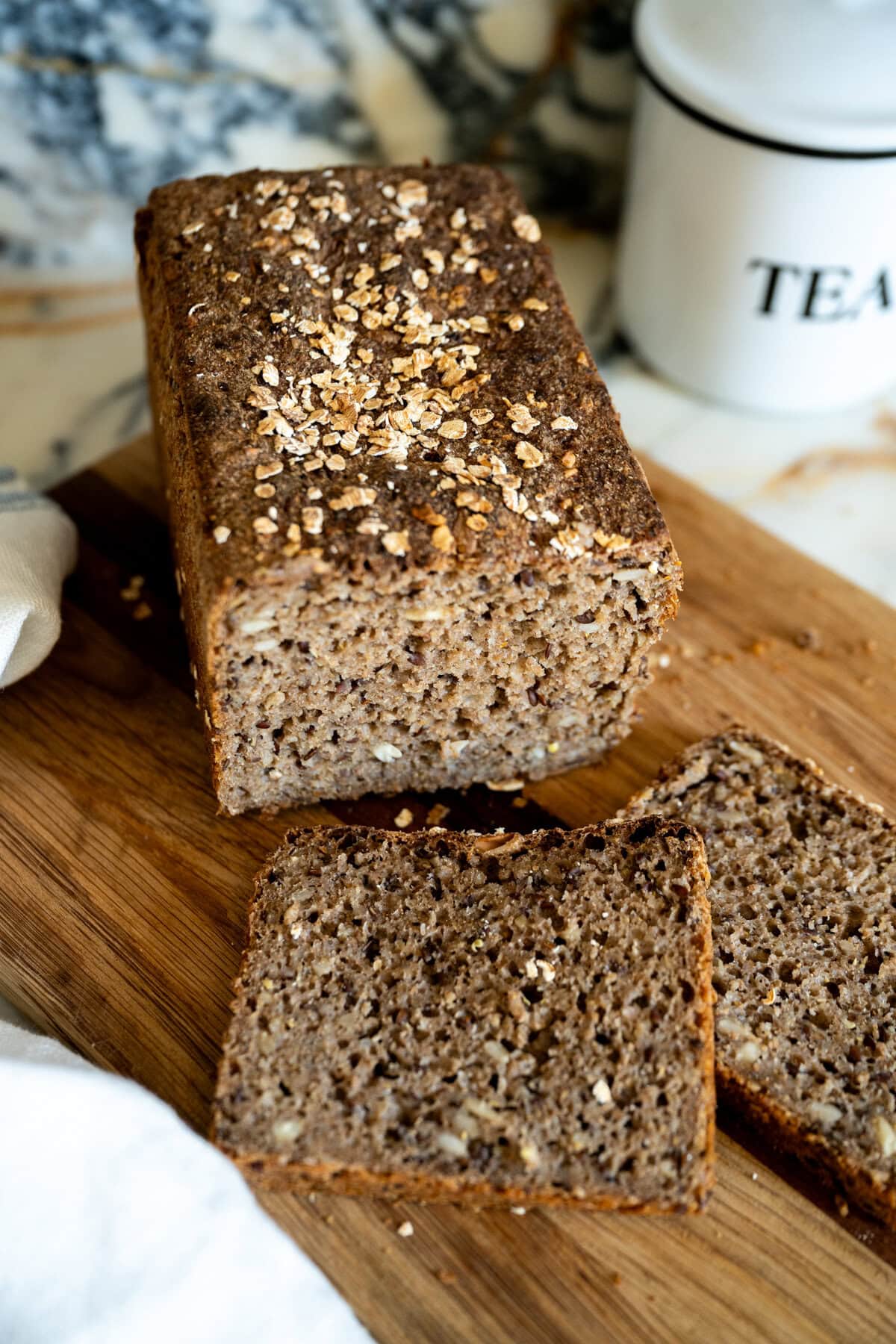 A loaf of dense whole grain sourdough Danish rye bread topped with oats sits on a wooden cutting board, with two slices cut in front of it. A white container labeled "TEA" is in the background.