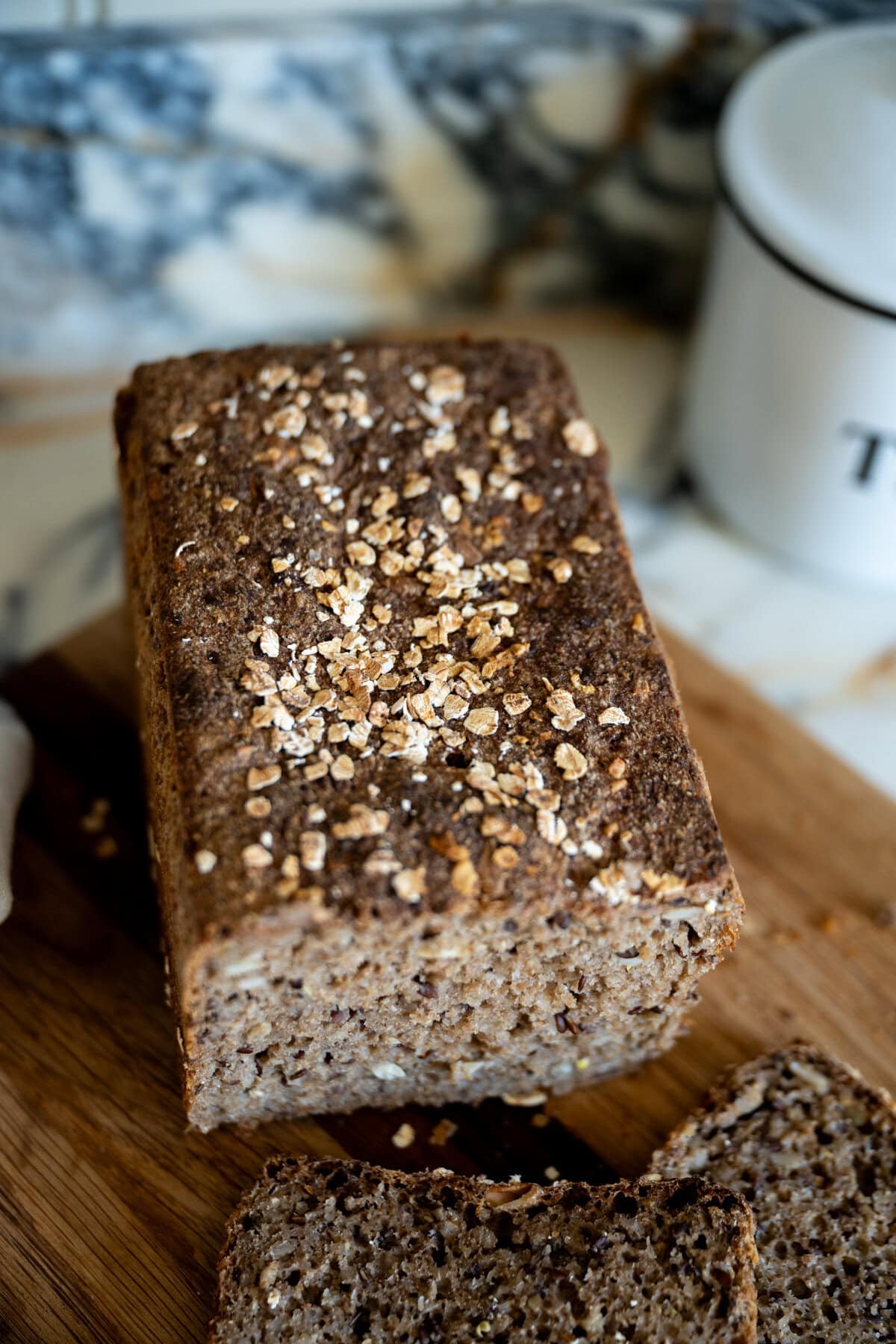 A loaf of whole grain sourdough Danish rye bread topped with oats sits on a wooden cutting board, with a few slices cut. A white container labeled “T” is in the background on a marble surface.