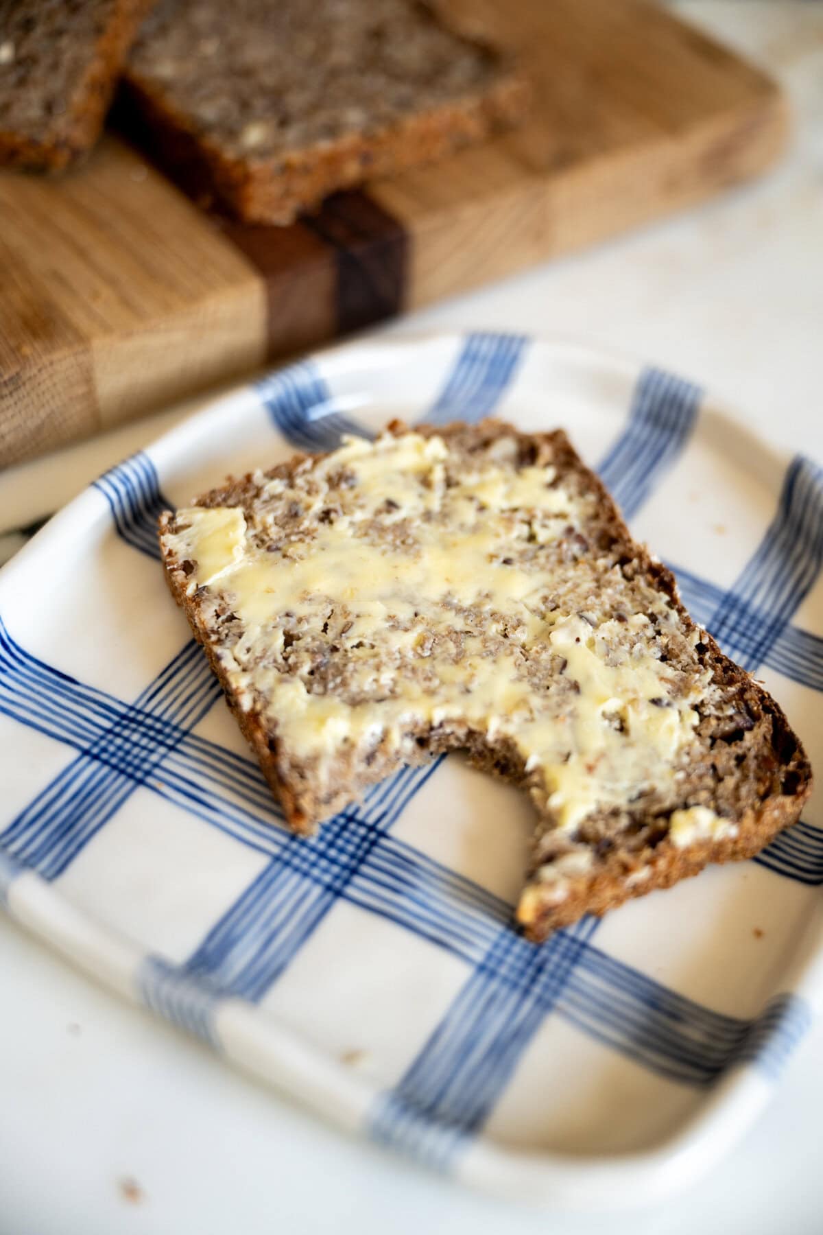 A buttered slice of homemade rugbrød Danish rye bread with a bite taken out, placed on a white plate with blue stripes. A wooden cutting board with more bread slices is in the background.