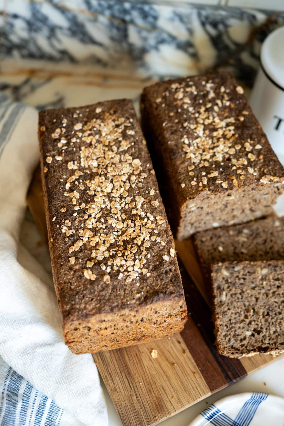 Two rectangular loaves of homemade sourdough Danish rye bread rugbrød topped with oats sit on a wooden cutting board, with a few slices cut from one loaf. A white towel and patterned background are also visible.