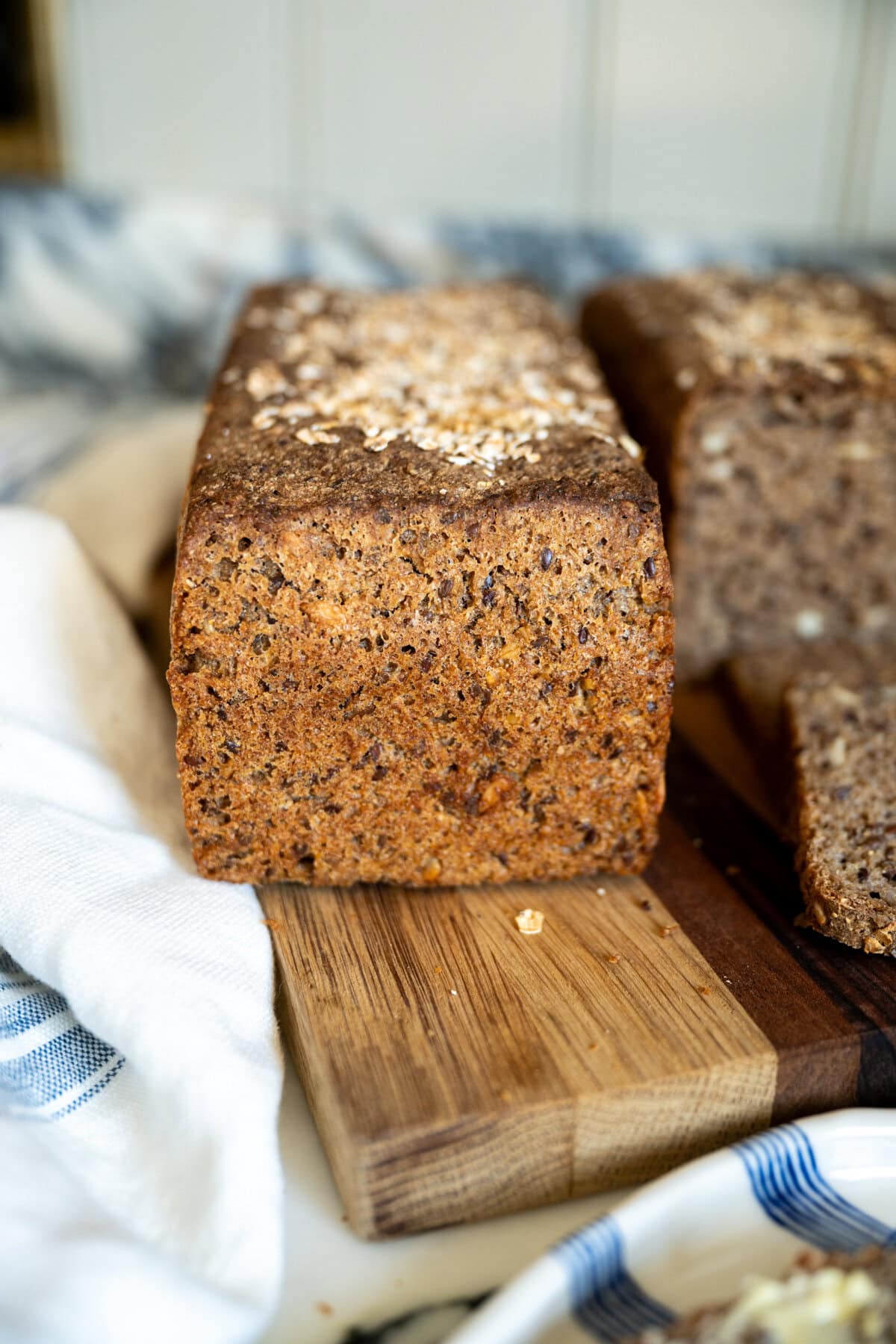 A close-up of a rectangular loaf of brown, whole-grain Danish rye rugbrød bread on a wooden cutting board, with a white cloth nearby. The bread has a textured crust and is topped with oats. Another loaf is partially visible in the background.