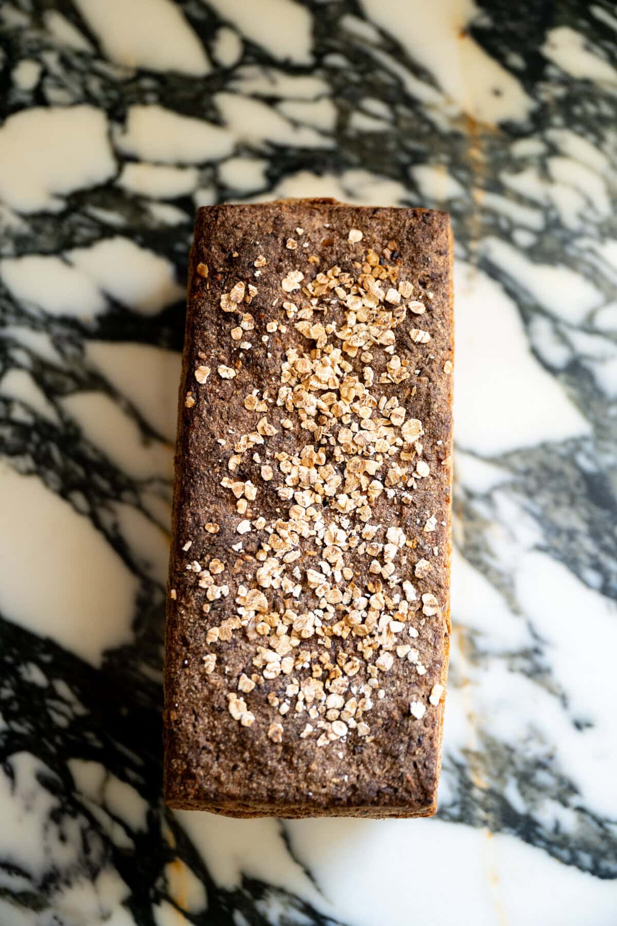 A rectangular loaf of Danish rye bread rugbrød with dark crust fully baked topped with sprinkled oats sits on a black-and-white marbled surface, viewed from above.