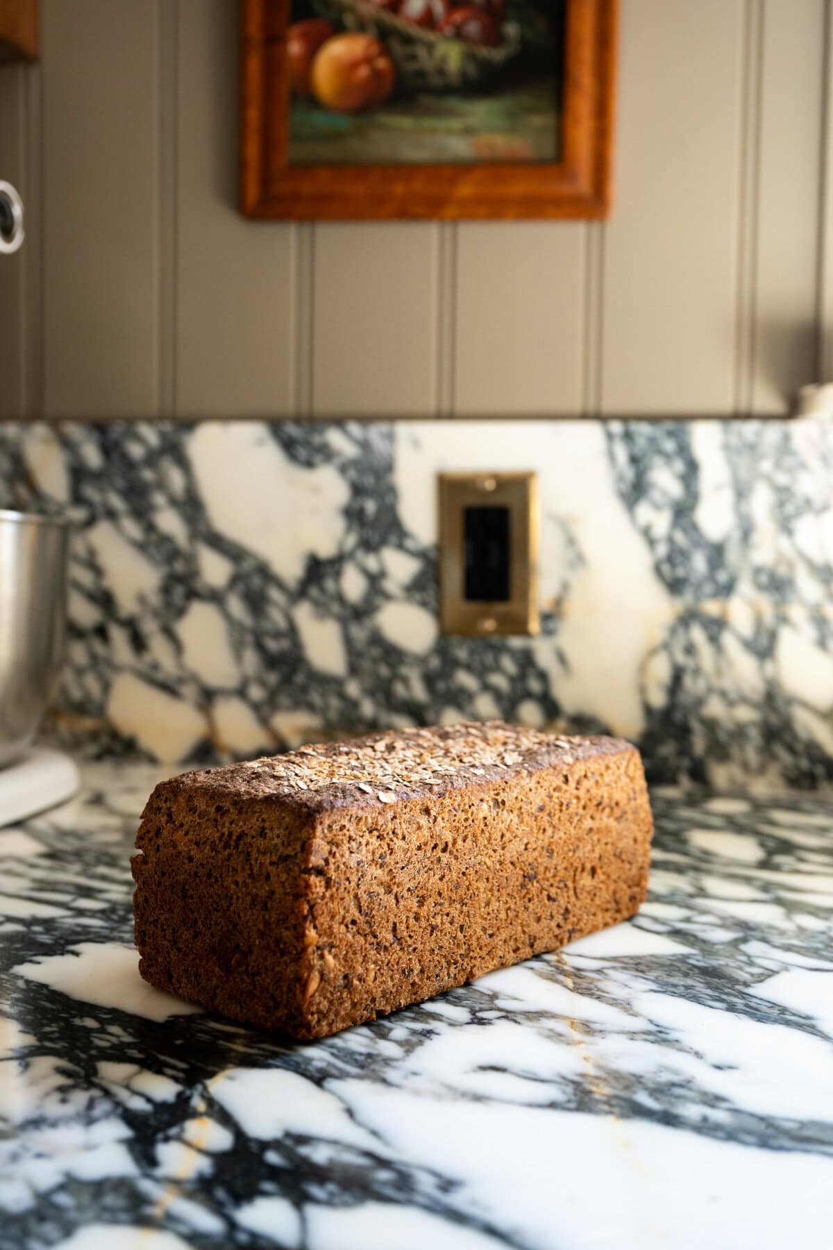 A rectangular loaf of dark, seeded Danish rye bread sits on a black-and-white marble countertop in a kitchen, with a painting of fruit hanging on the wall in the background.