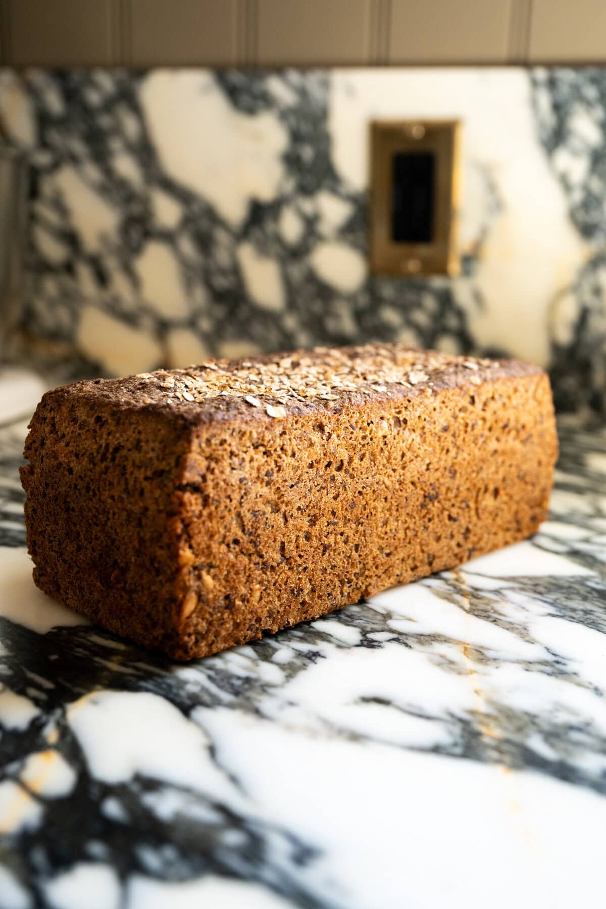 A loaf of dark, whole grain sourdough Danish rye bread with a textured crust sits on a marbled countertop in a kitchen.