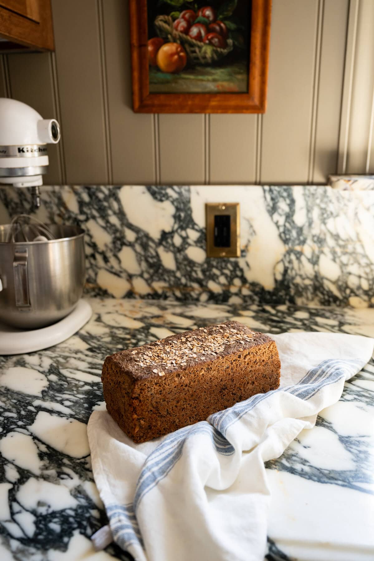 A loaf of homemade whole grain Danish rye bread rugbrød topped with oats rests on a white and blue striped cloth on a marble countertop. A stand mixer and a framed fruit painting are visible in the background.