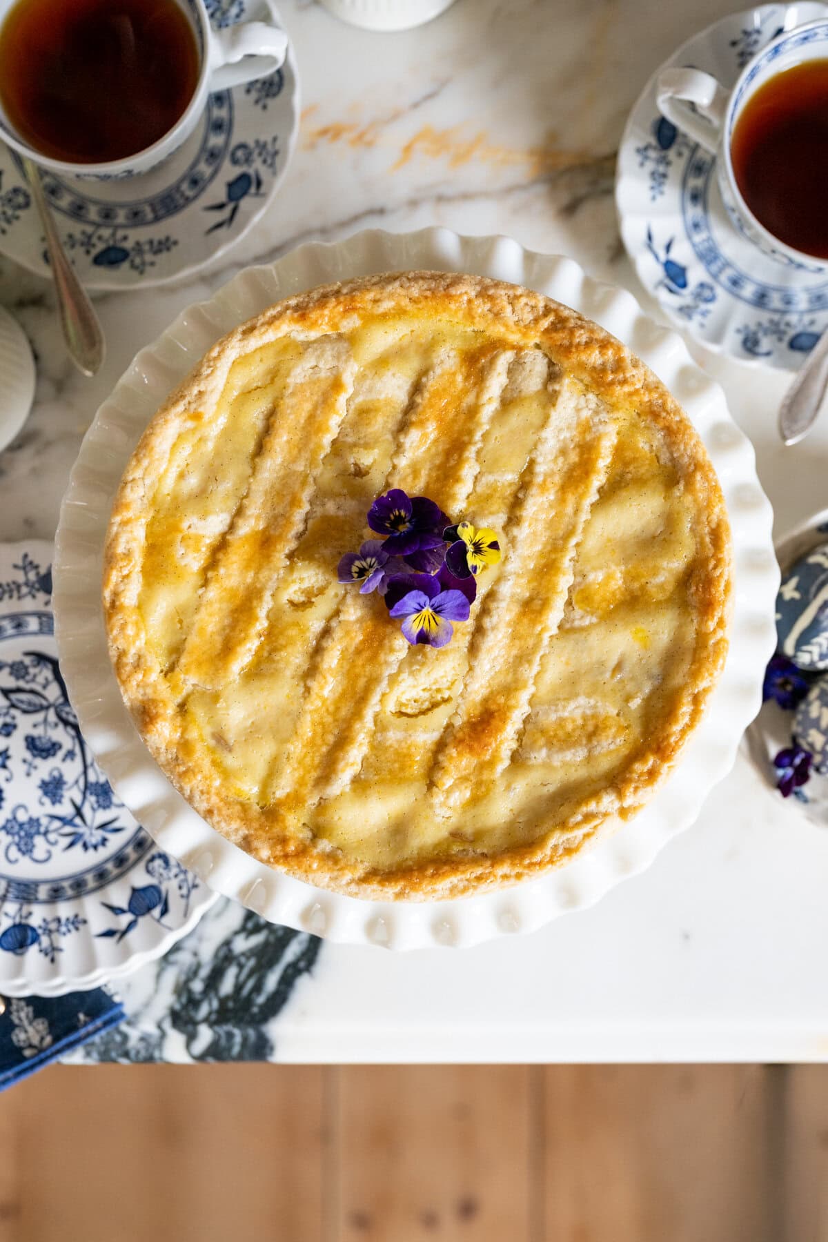 A Pastiera Napoletana with a lattice top is garnished with purple and yellow edible flowers, sitting on a white plate surrounded by blue and white patterned teacups filled with tea on a marble table.