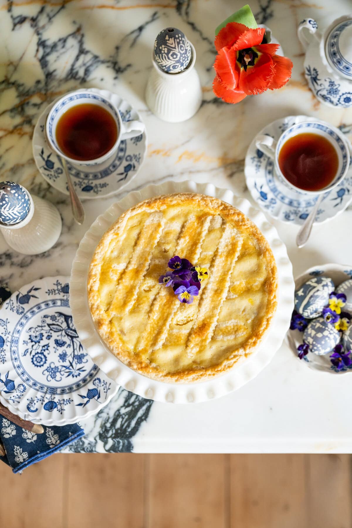 A round Pastiera Napoletana pie topped with edible flowers sits on a marble table, surrounded by blue-and-white teacups filled with tea, saucers, a flower vase with a red bloom, and a set of salt and pepper shakers.