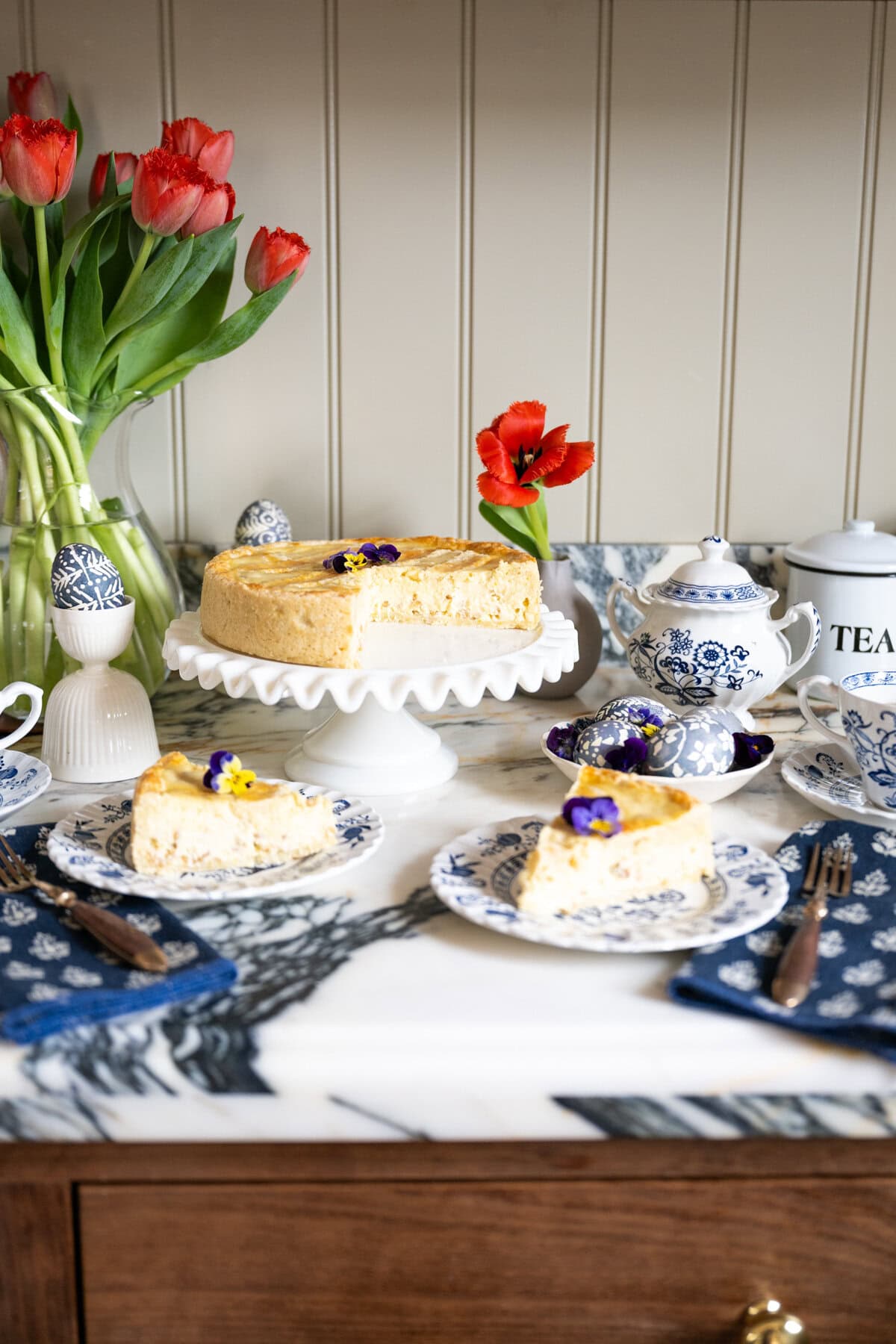 A traditional Easter Pastiera Napoletana pie garnished with edible flowers sits on a white cake stand, with two slices on plates beside blue-and-white teacups. Red tulips, napkins, and a tea canister complete the elegant table setting.