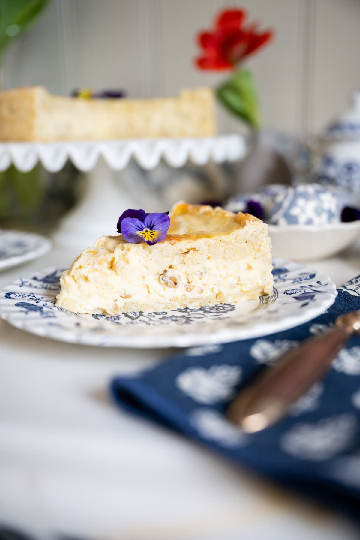 A slice of Pastiera Napoletana or Italian Easter grain pie garnished with a purple edible flower sits on a decorative blue and white plate, with a cake on a stand and a red flower in the blurred background. A napkin and utensil are nearby.