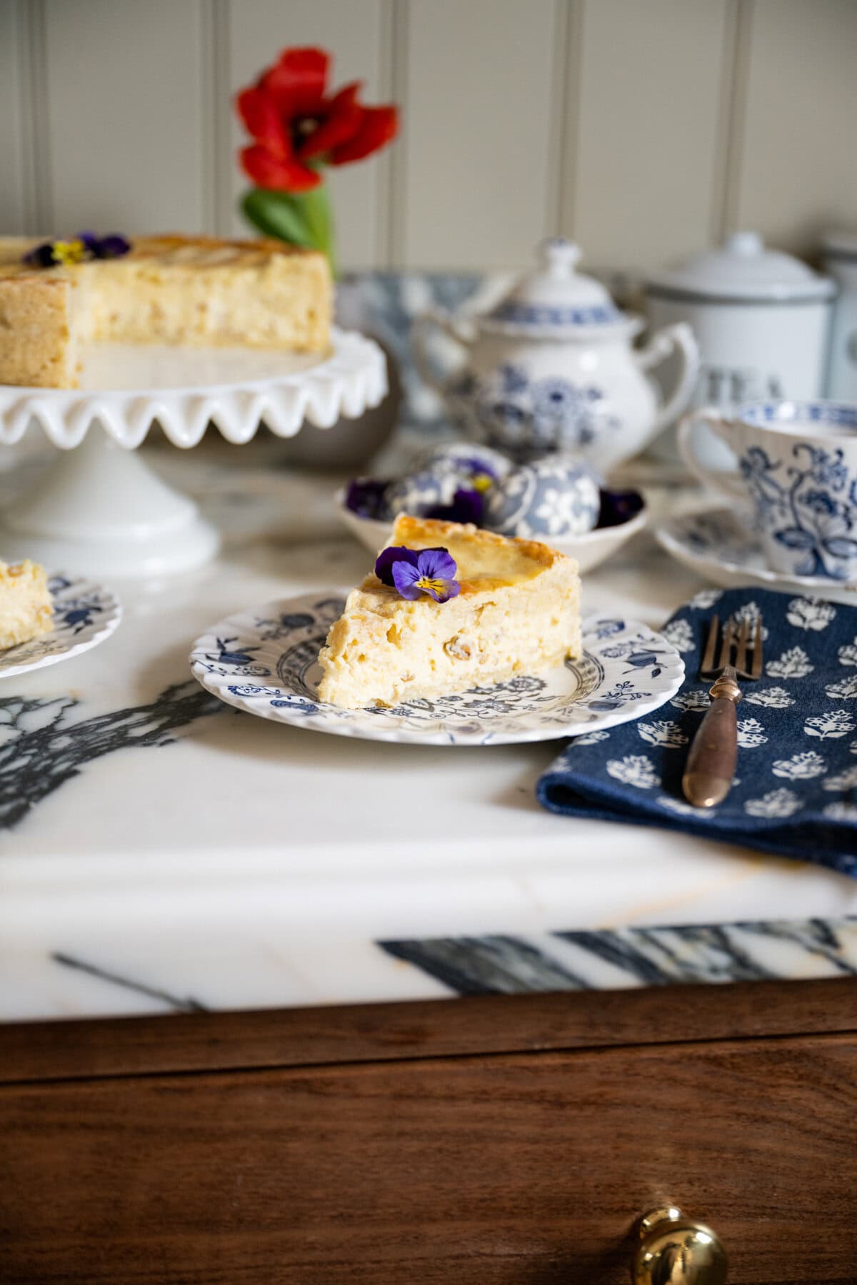 A slice of traditional Pastiera Napoletana topped with a purple flower sits on a decorative plate, next to a fork and a blue floral napkin. In the background, there is a cake stand, teapot, teacup, and a red flower in a vase.
