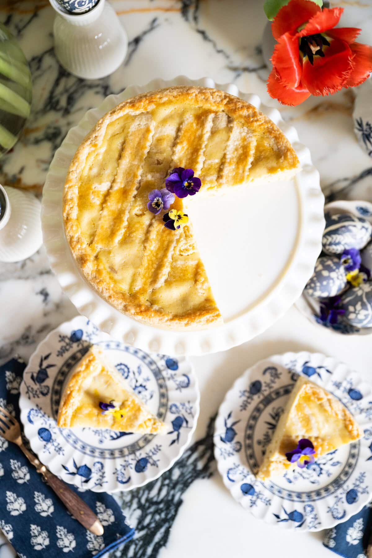 A round Pastiera Napoletana Easter pie with a lattice top sits on a white cake stand, with one slice removed and placed on a blue-and-white floral plate. Two plates with pie slices are decorated with edible flowers. A red flower and teacup are nearby.