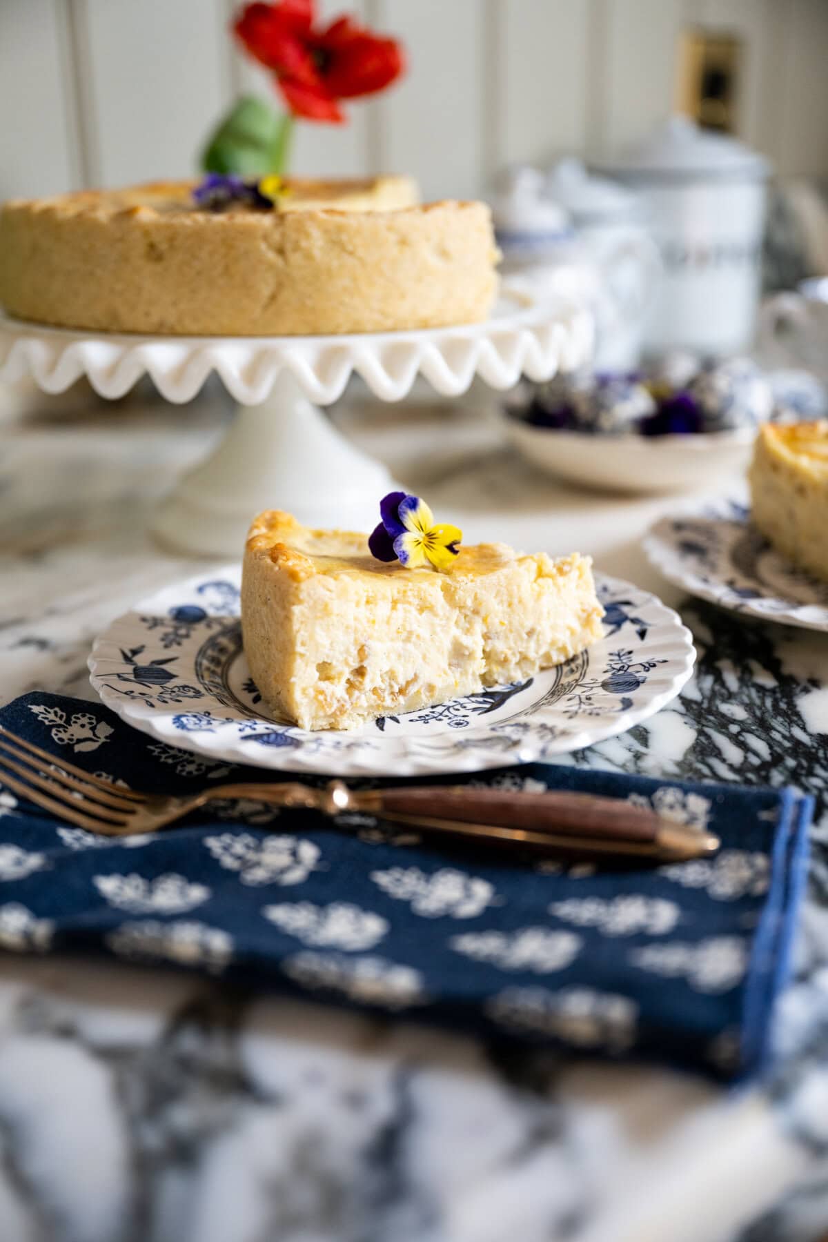 A slice of Pastiera Napoletana, a traditional Italian Easter ricotta and grain pie, garnished with an edible flower sits on a blue and white plate, with the rest of the pastiera on a cake stand in the background, atop a marble table.