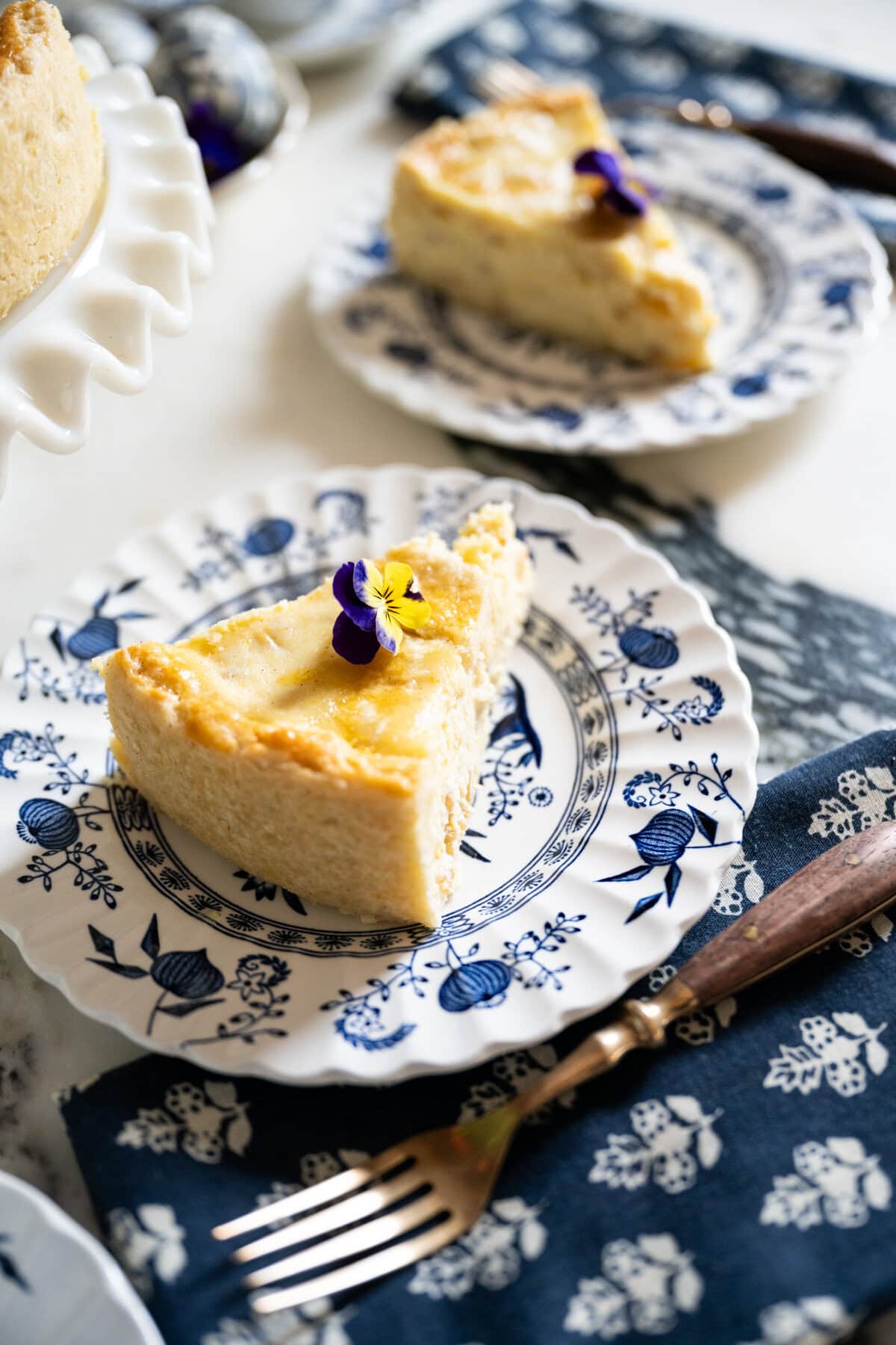 A slice of traditional Pastiera Napoletana garnished with a purple and yellow edible flower is served on a decorative blue and white plate, next to a blue patterned napkin and a fork. Another slice is in the background.