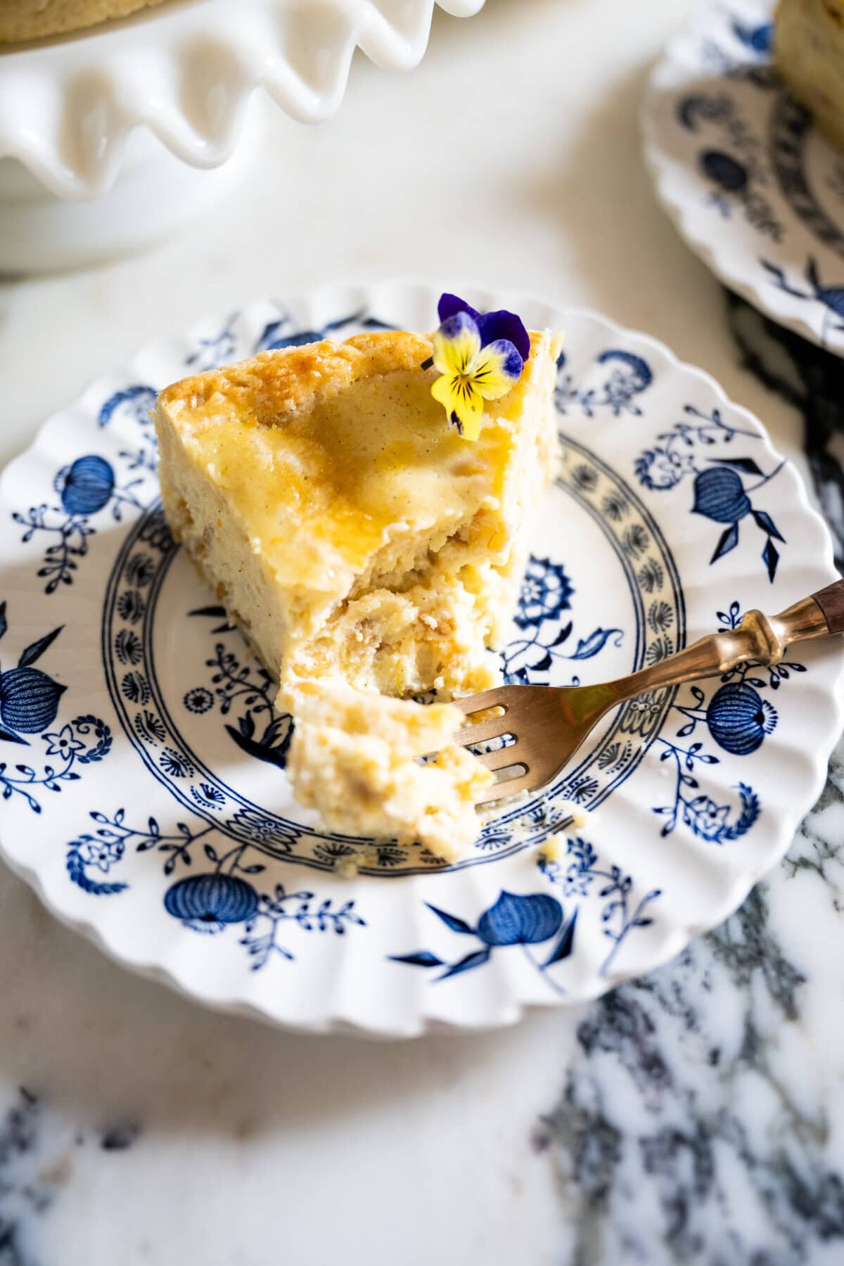 A slice of creamy Pastiera Napoletana garnished with a purple and yellow edible flower sits on a blue-and-white floral plate, with a fork holding a bite-sized piece on a marble surface.