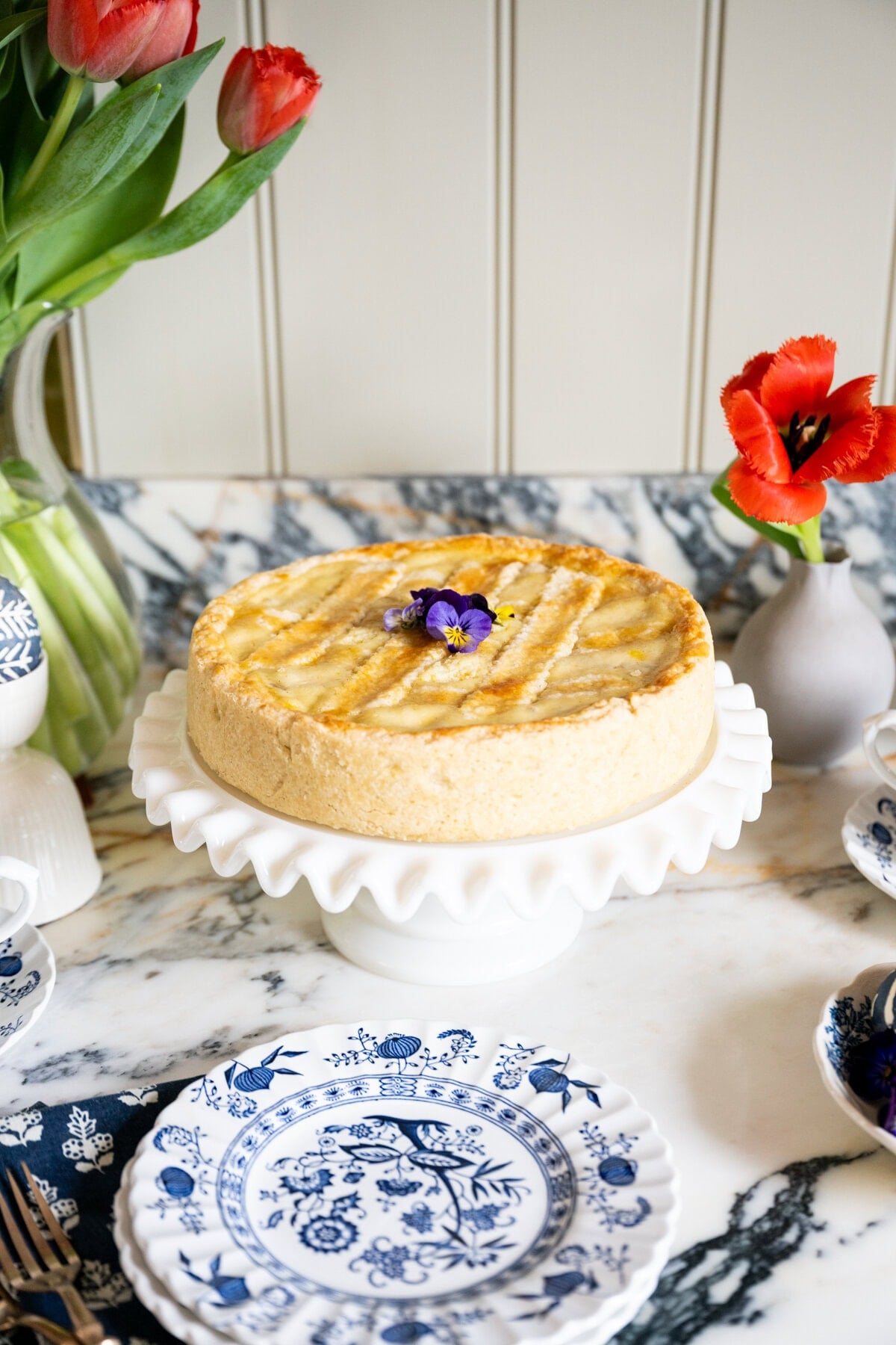 A round Pastiera Napoletana with a lattice top, decorated with edible flowers, sits on a white cake stand, surrounded by blue and white plates on a marble table. Red tulips in vases brighten the background.