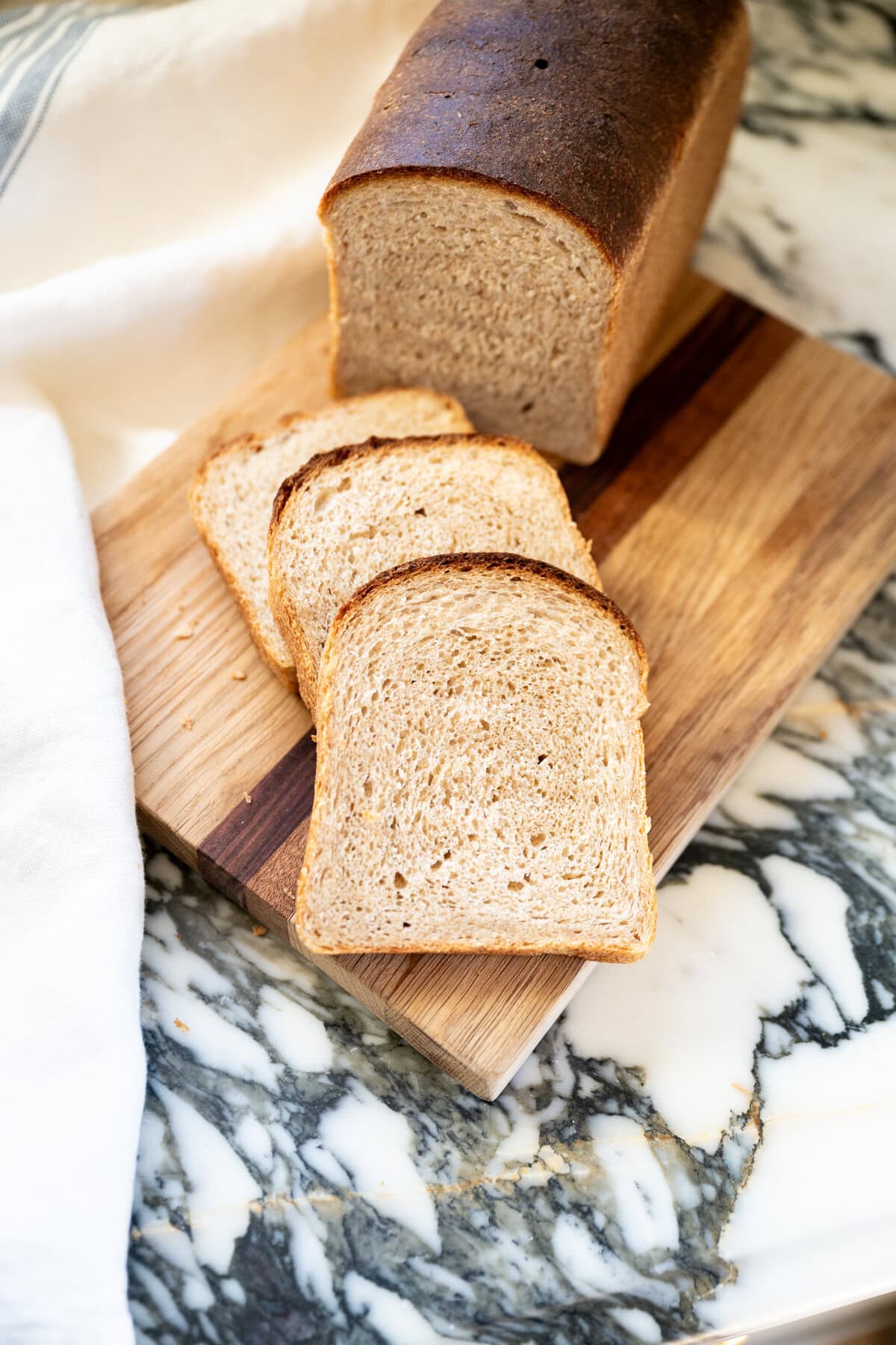 A loaf of soft honey whole wheat sourdough bread with a dark crust sits on a wooden cutting board, with three slices cut and fanned out. The board is on a marble countertop next to a white cloth.