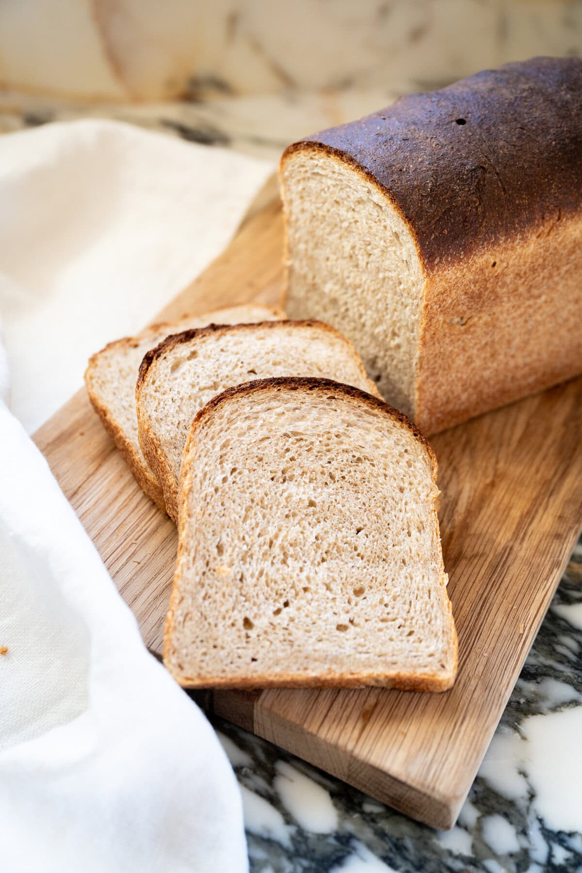 A loaf of whole wheat sourdough sandwich bread with a dark, golden-brown crust sits on a wooden cutting board. Three slices are cut and fanned out in front of the loaf. A white cloth is placed to the side.