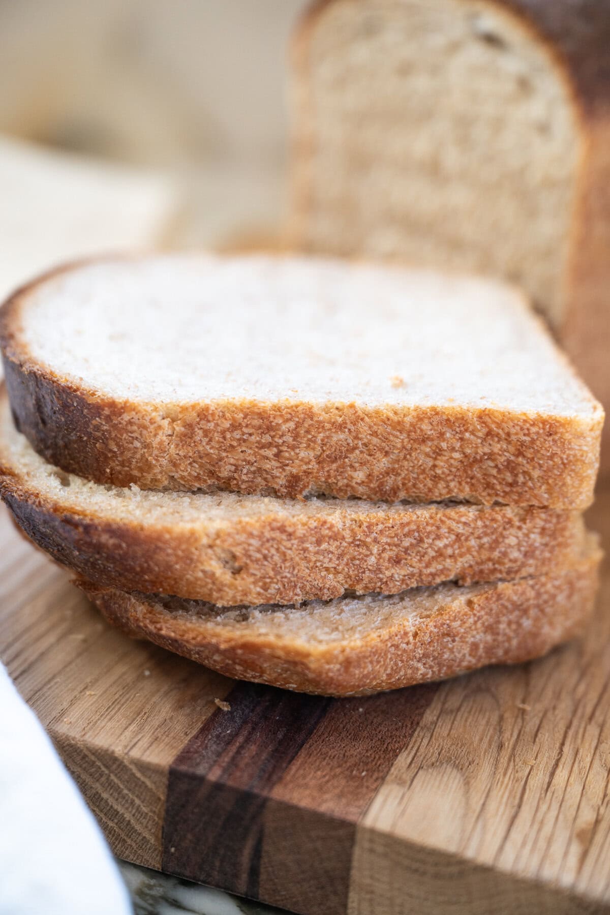 Three slices of soft whole wheat sourdough sandwich bread stacked on a wooden cutting board, with a loaf of bread in the background. The bread appears fresh and slightly textured, with a light, soft interior and golden-brown crust.