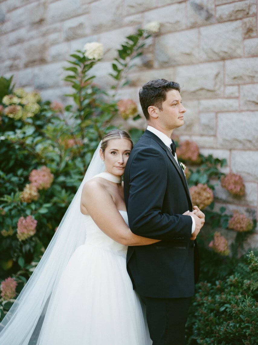 A bride in a white gown and veil embraces a groom in a black suit as they stand before a stone wall and lush greenery, their calm serenity beautifully captured by a Nantucket photographer.