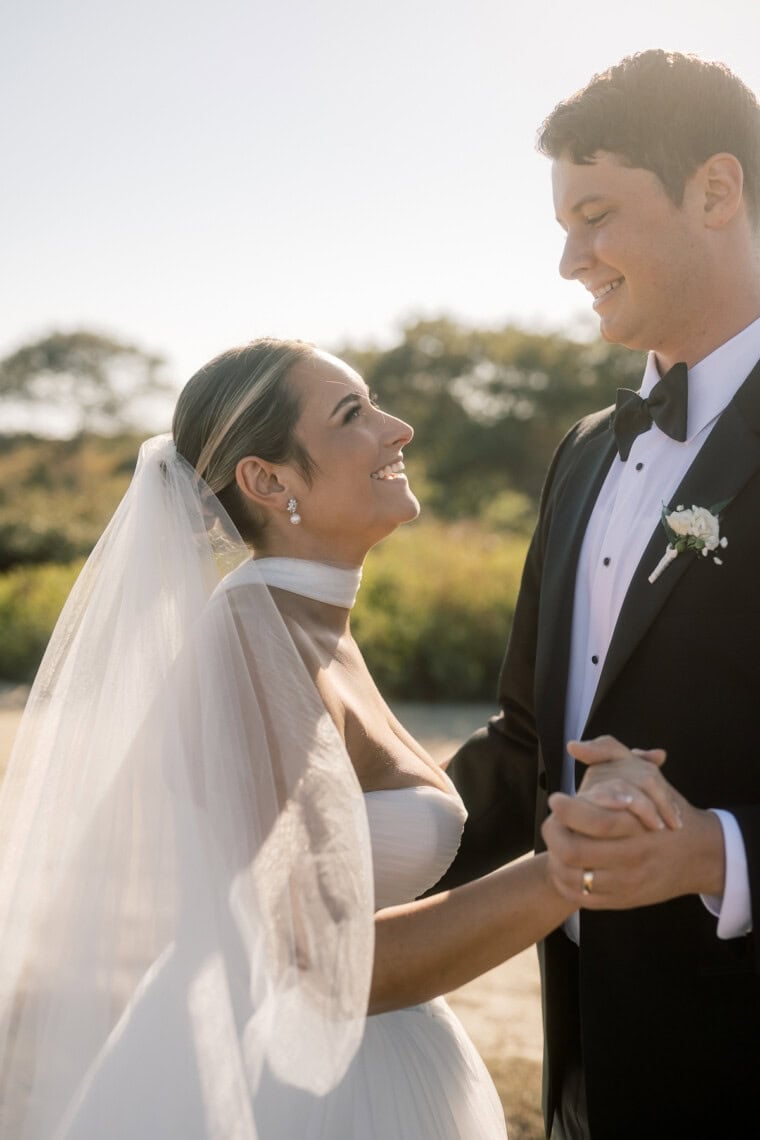 A bride in a white dress and veil smiles up at a groom in a black tuxedo as they hold hands outdoors on a sunny day, greenery and trees softly blurred behind them—captured beautifully by a Nantucket photographer.