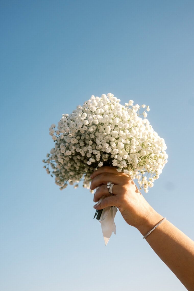 A hand holding a bouquet of small white flowers against a clear blue sky, adorned with rings and a delicate bracelet—captured in the signature airy style of a Nantucket photographer.